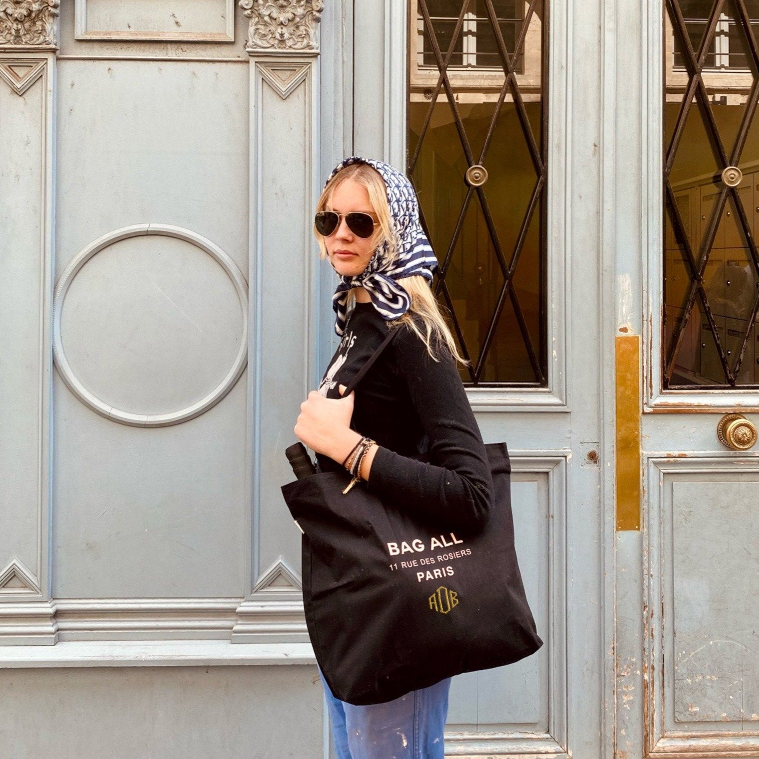 Bag-all Paris Le Marais Tote with Zipper, stylish black cotton tote bag featuring white Le Marais address print, side view worn by person in casual outfit near Parisian doorway