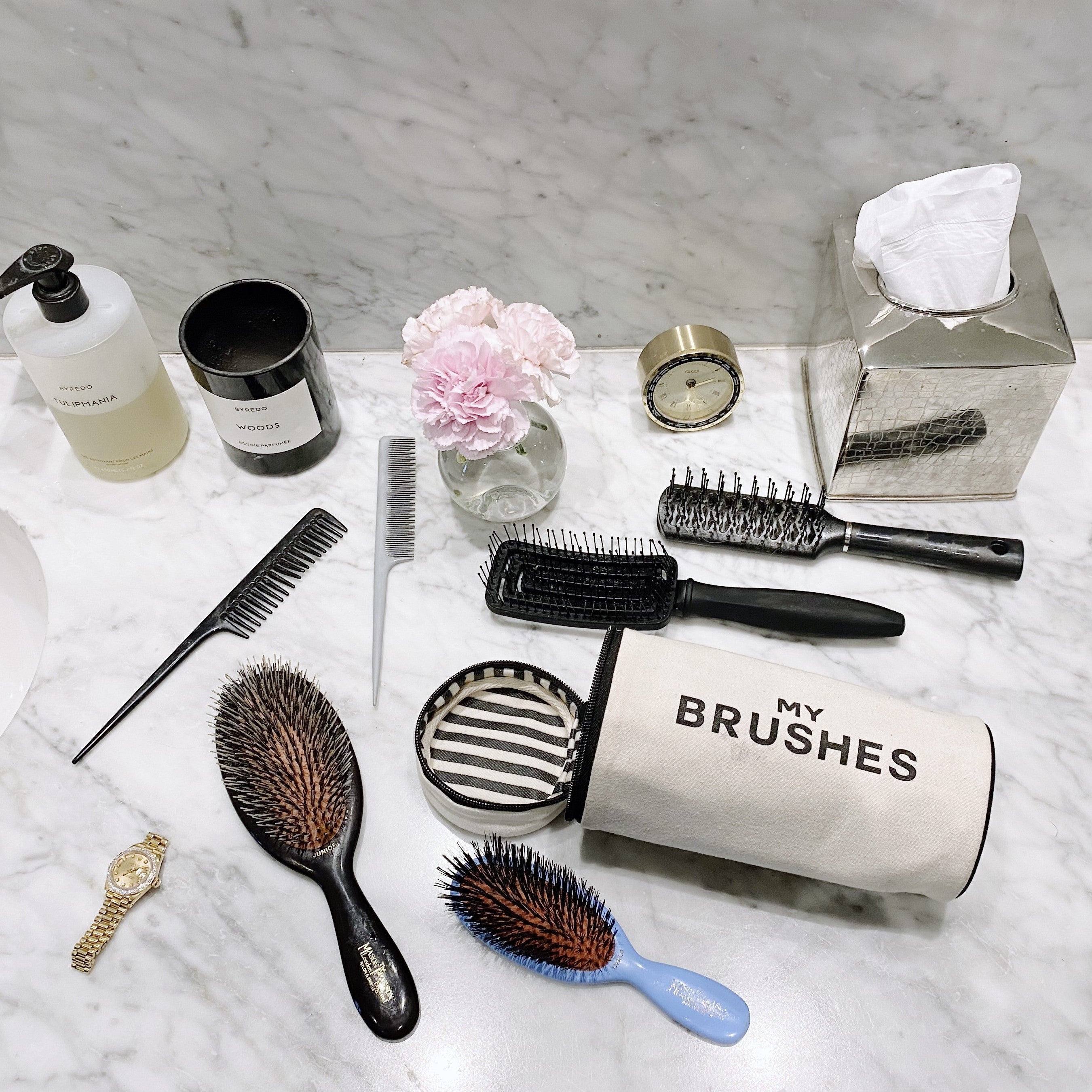 Bag-all My Brushes organizer with stylish hair care essentials including brushes, combs, and toiletries displayed on elegant marble surface with pink carnation accent
