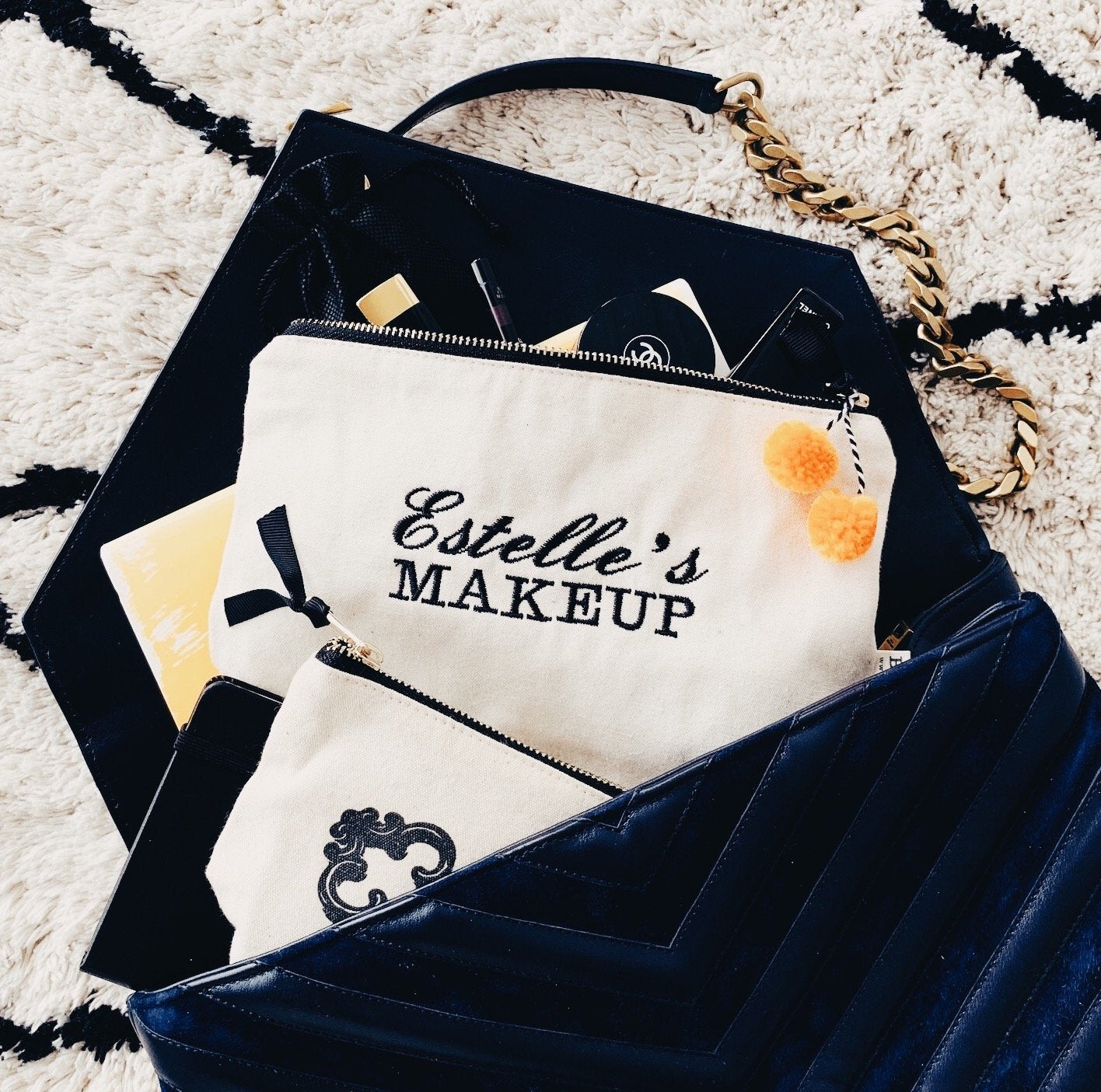 Bag-all personalized cotton makeup bag with embroidered name text. Stylish black and white design with zipper closure and classic chain detail on plush carpet.