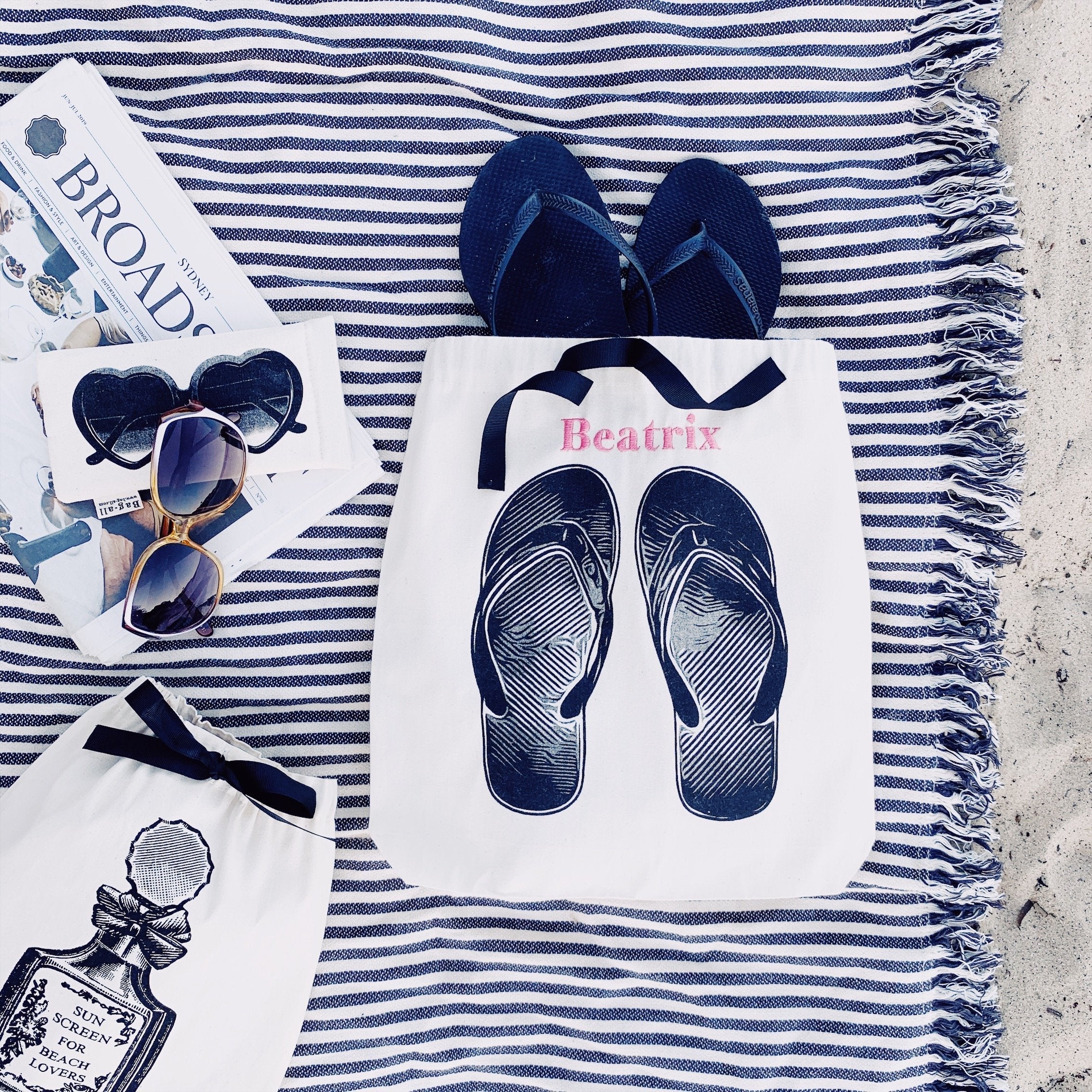 Bag-all Cream Flip Flops Shoe Bag featuring navy blue flip flop design on white cotton, displayed on striped beach blanket with sunglasses and beach accessories