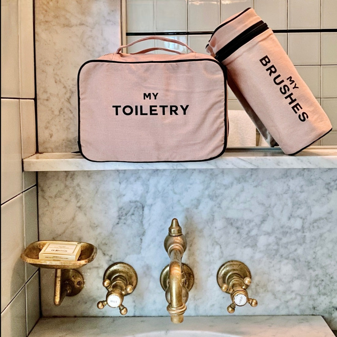 Bag-all pink cotton My Toiletry travel case and My Brushes makeup organizer featuring black text, displayed on marble bathroom shelf with vintage gold fixtures
