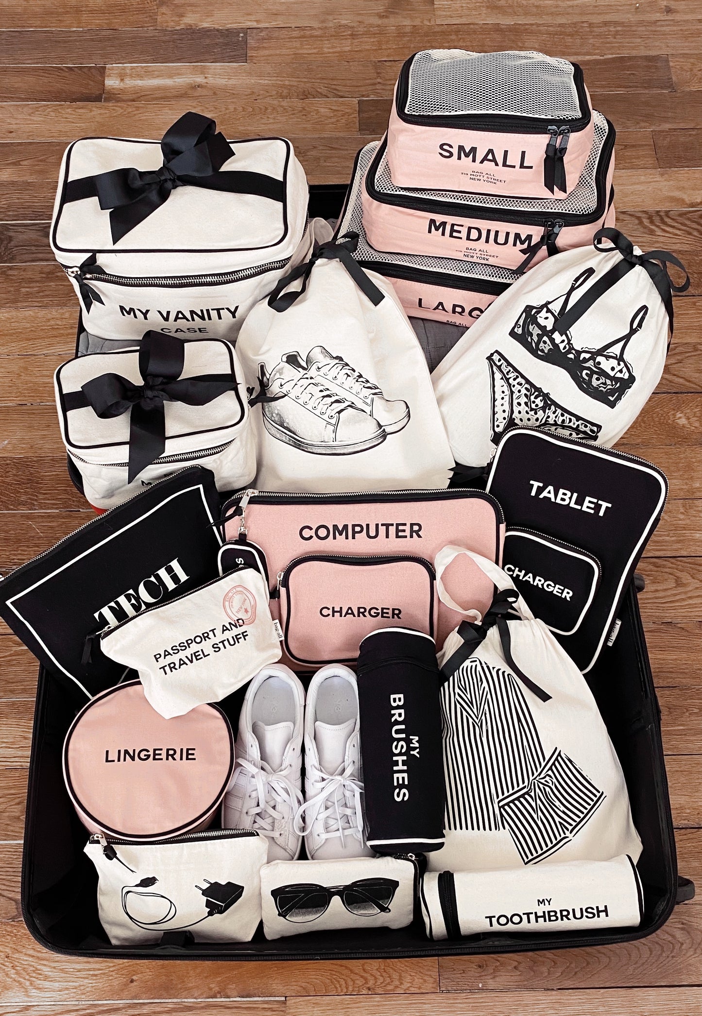 Bag-all black and pink travel organization cases including Toothbrush Travel Case, displayed on wooden floor with tech pouches, lingerie bags, and shoe organizers for efficient packing