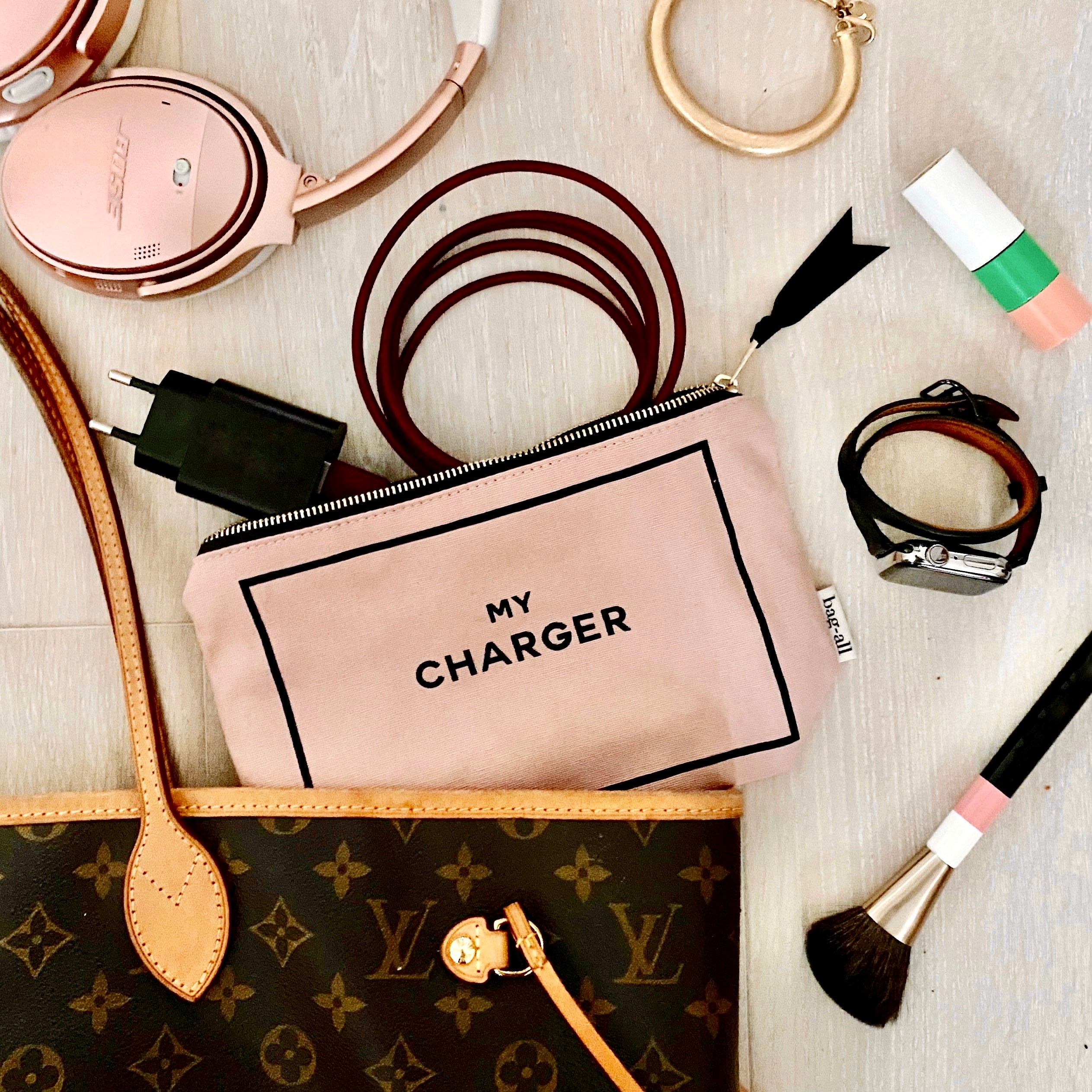 Bag-all Charger Pouch in blush pink with black text, shown with chargers, cords, and accessories against wooden surface, next to designer handbag for size reference