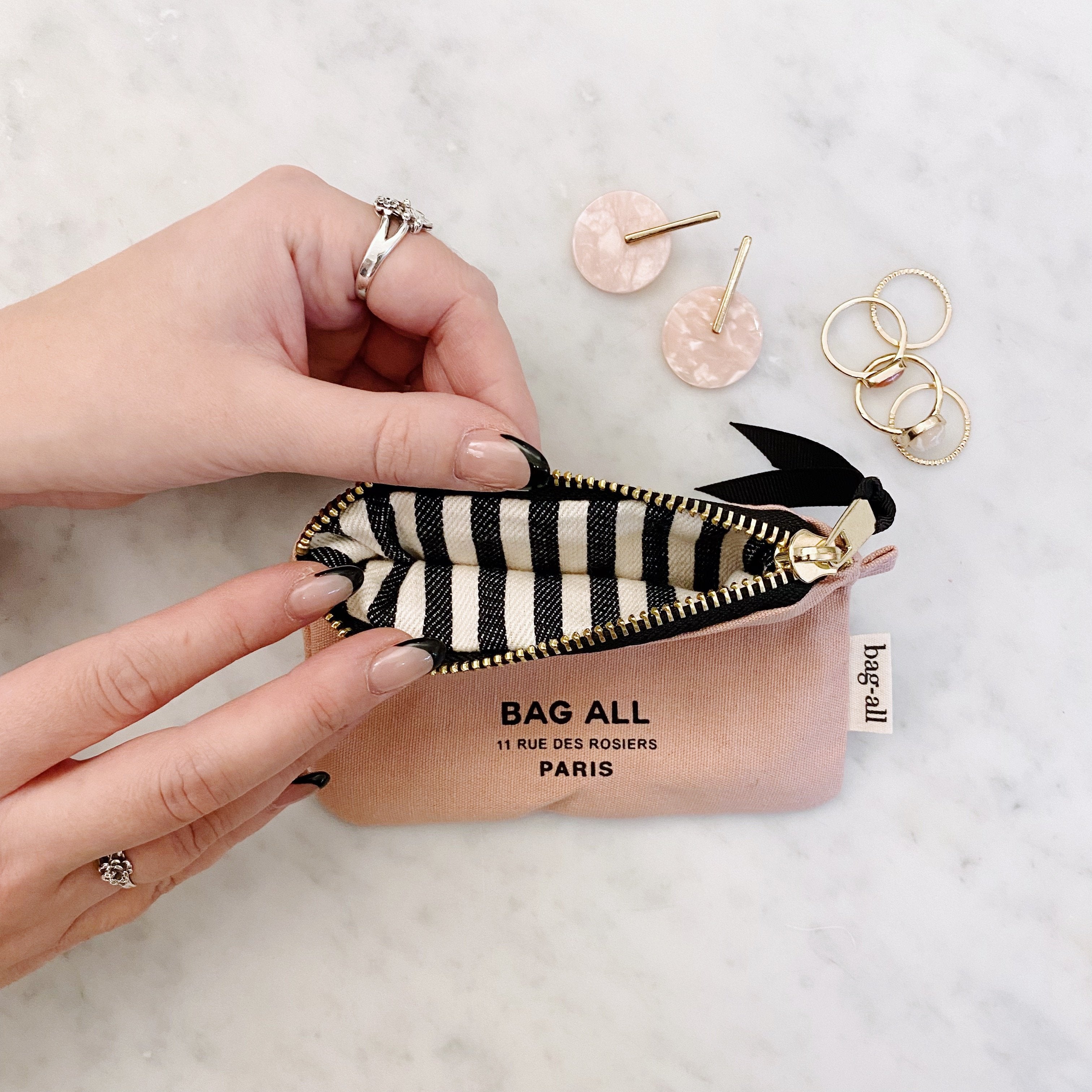 Bag-all Mini Trinket Pouch in pink/blush with black and white striped interior, gold metal zipper, displayed with jewelry pieces on marble surface, showcasing compact storage solution