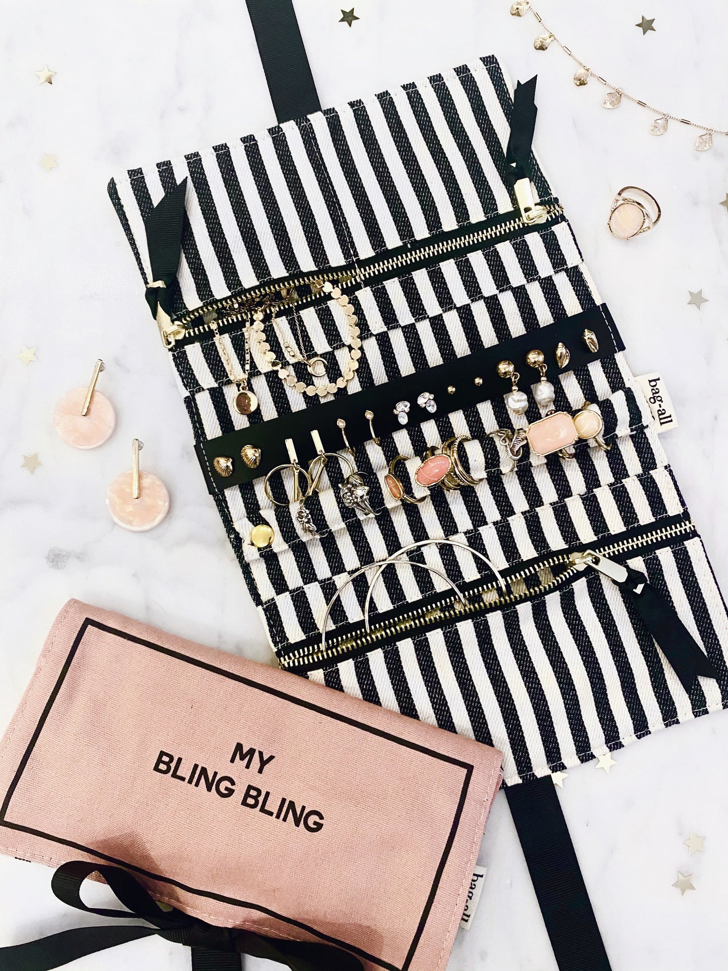 Bag-all Jewelry Organizer Travel Pouch in pink blush with black and white striped interior, featuring multiple compartments, earring holder, and ring storage, showcasing organized jewelry display against decorative background