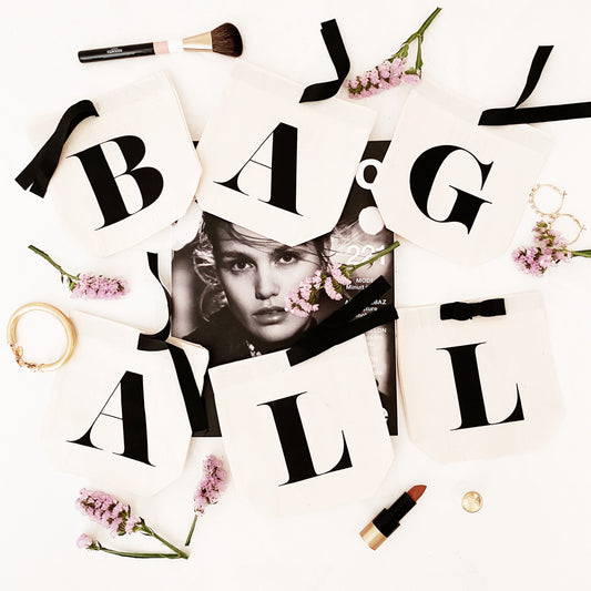 Bag-all Small Letter Bags Classic in cream with stylized typography, arranged with pink flowers, makeup brushes and lipstick on white background, perfect for organizing essentials
