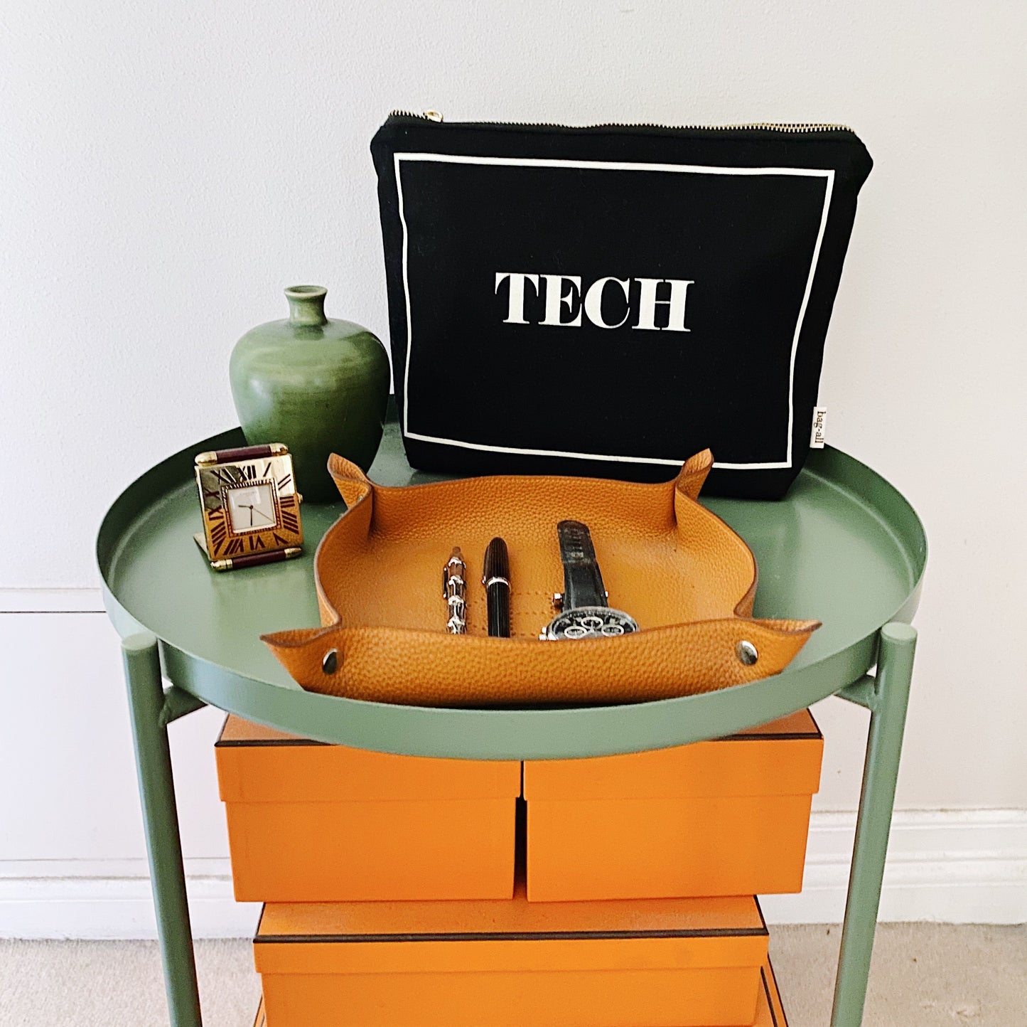 Bag-all Tech Pouch in black with white border, displayed on green side table with leather catchall tray and decorative vase, showcasing modern tech organization solution