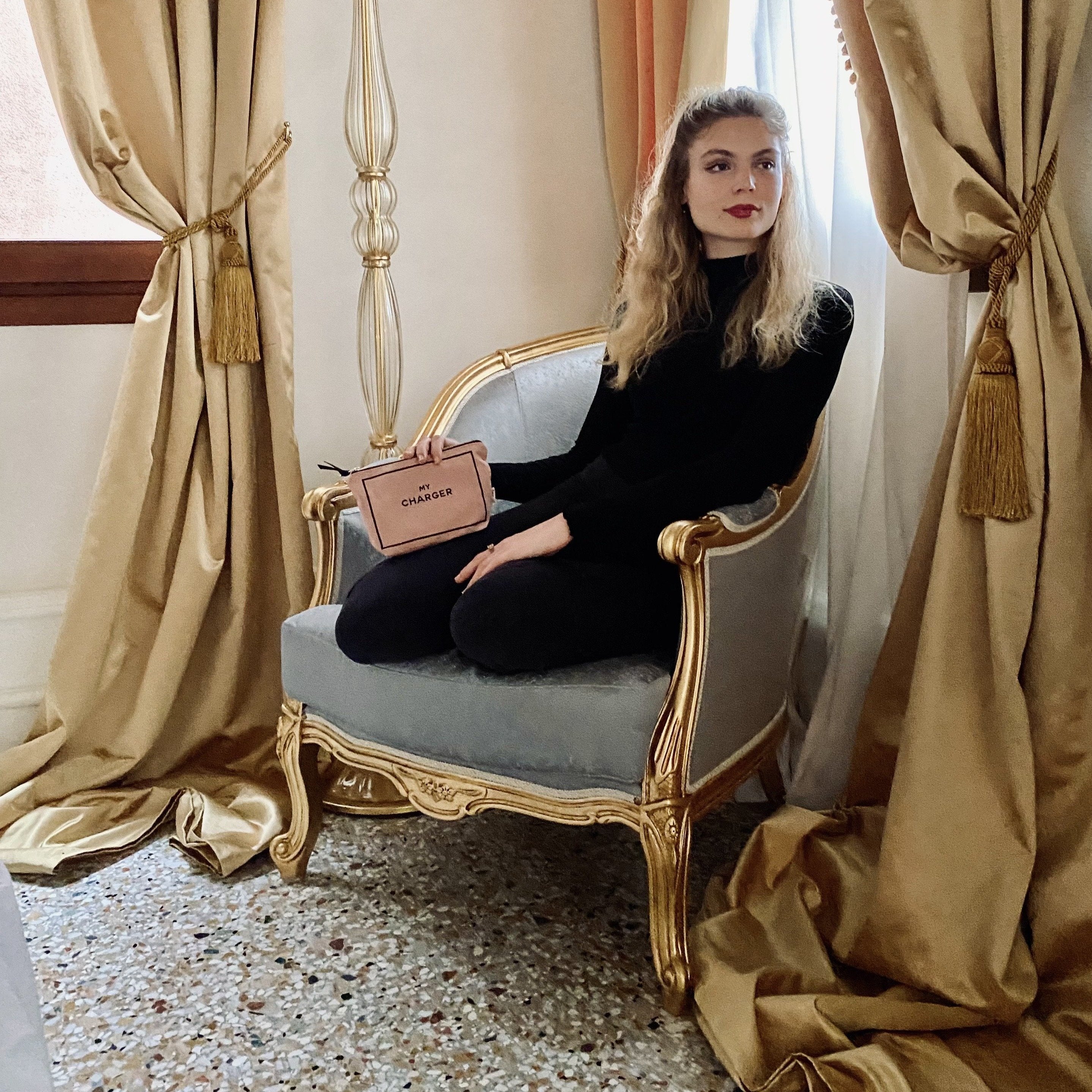 Bag-all Charger Pouch in blush pink shown in elegant setting with vintage chair, gold curtains and classic interior design, demonstrating luxury storage solution for tech accessories