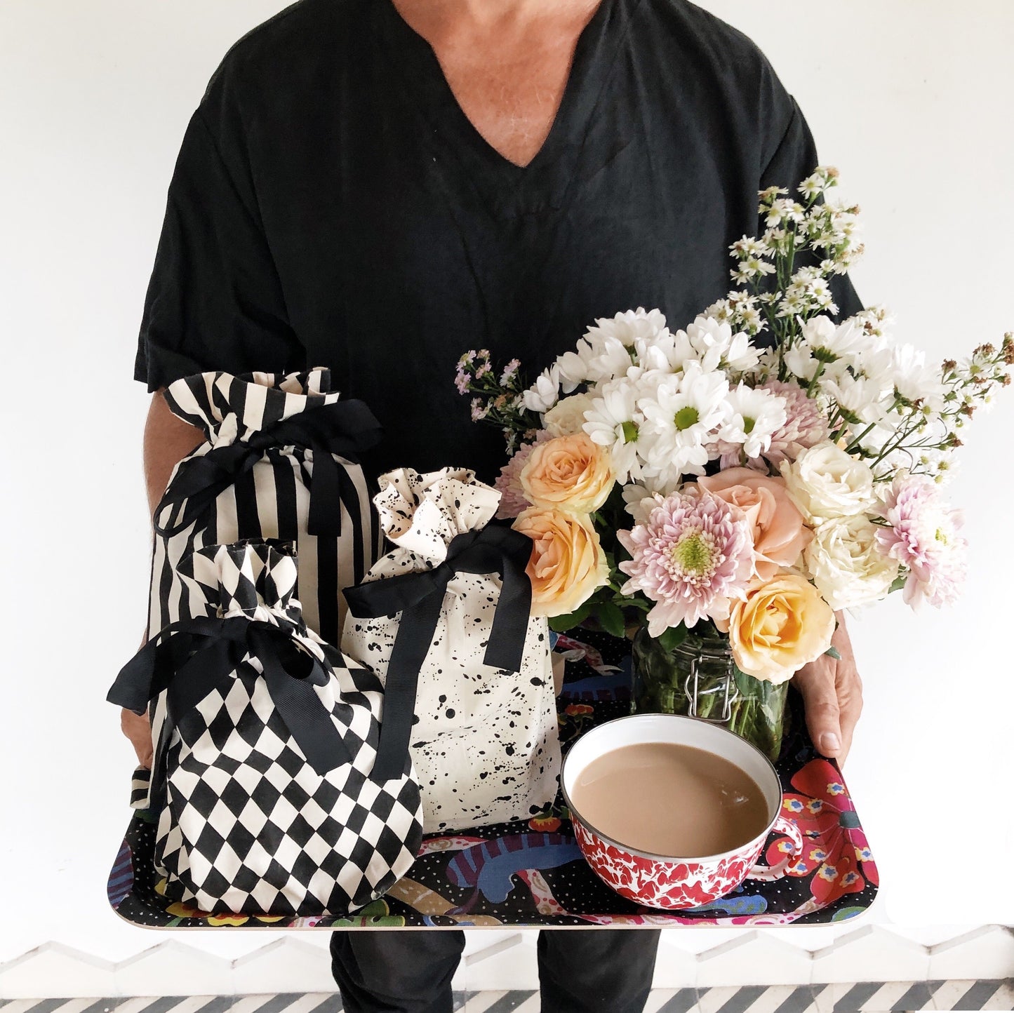 Bag-all Gift Bag Striped Medium shown with fresh flowers and coffee, featuring classic black and white stripes in reusable cotton, displayed alongside checkered patterns and floral arrangements