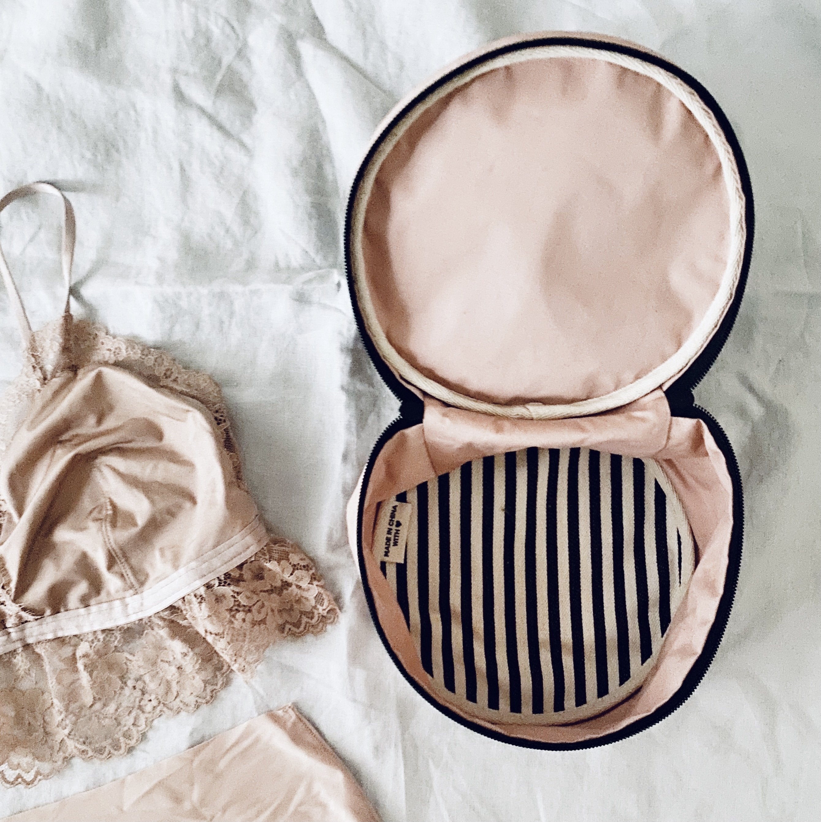 Bag-all Round Lingerie Case in blush pink with black striped interior lining, featuring cotton construction and metal zipper, shown with delicate lace undergarments on white fabric background