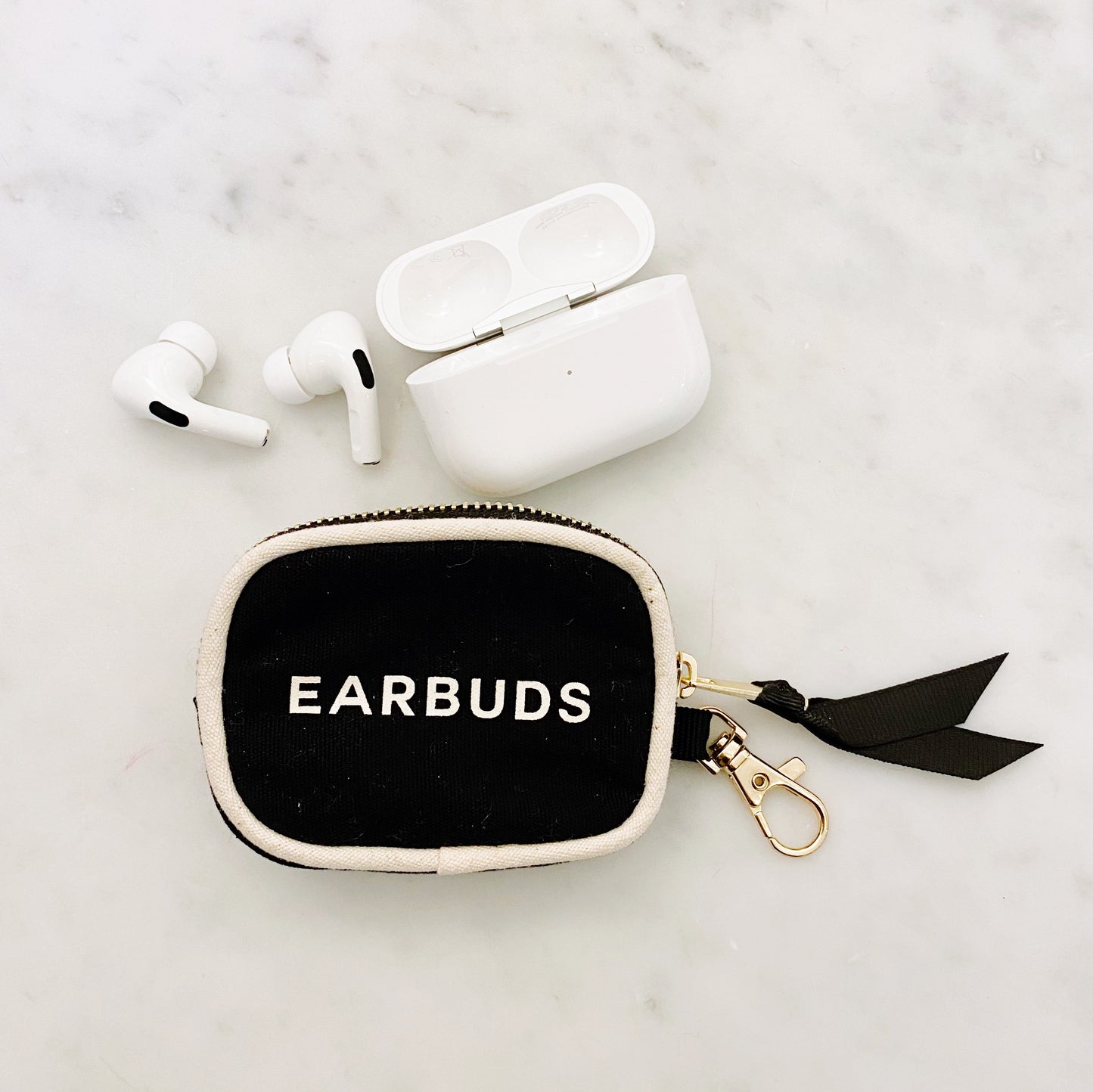 Bag-all Black Earbuds Case with gold clasp and zipper featuring white piping and "EARBUDS" text, shown with white wireless earphones on marble surface