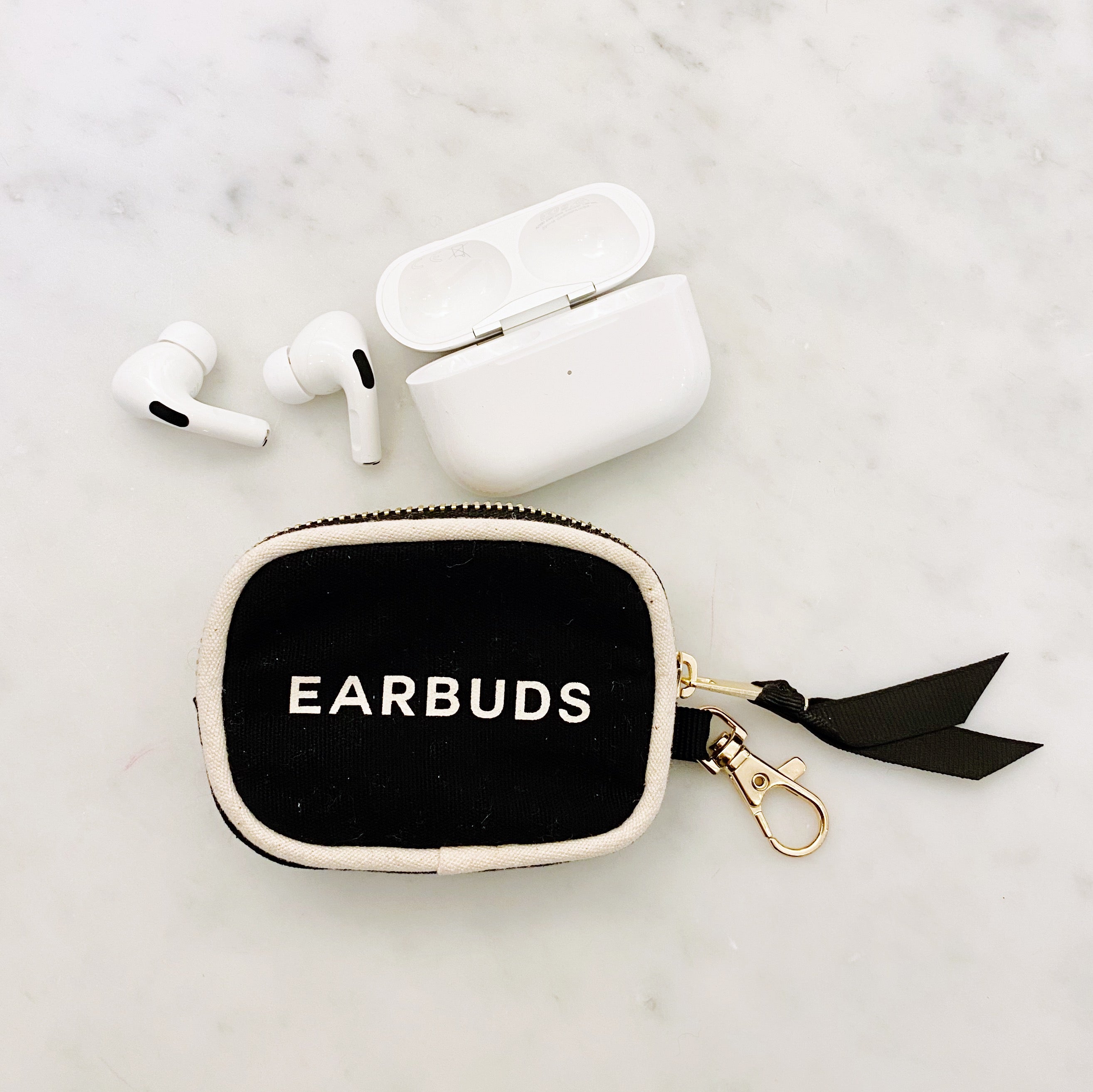 Bag-all Black Earbuds Case with gold clasp and zipper featuring white piping and "EARBUDS" text, shown with white wireless earphones on marble surface