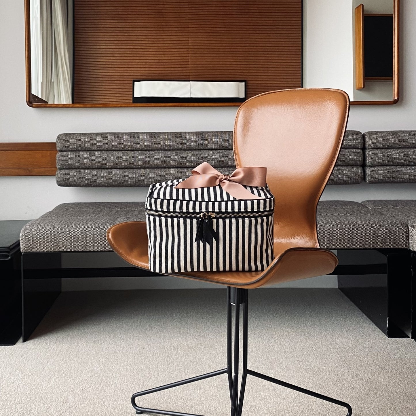 Bag-all Medium Box Makeup & Toiletry in black and white stripes with pink bow accent, displayed on brown leather chair, features gold zipper and waterproof lining for travel organization