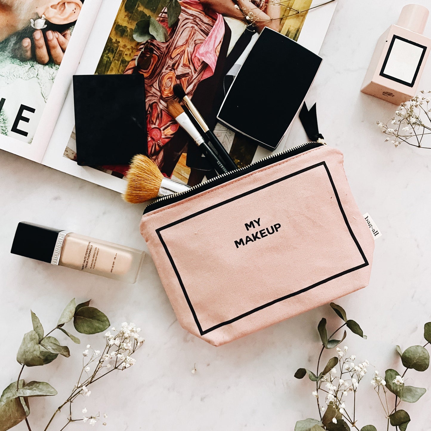 Bag-all My Makeup Pouch in blush pink canvas with black text and gold zipper, featuring coated interior lining, styled with makeup brushes and floral accents on white surface
