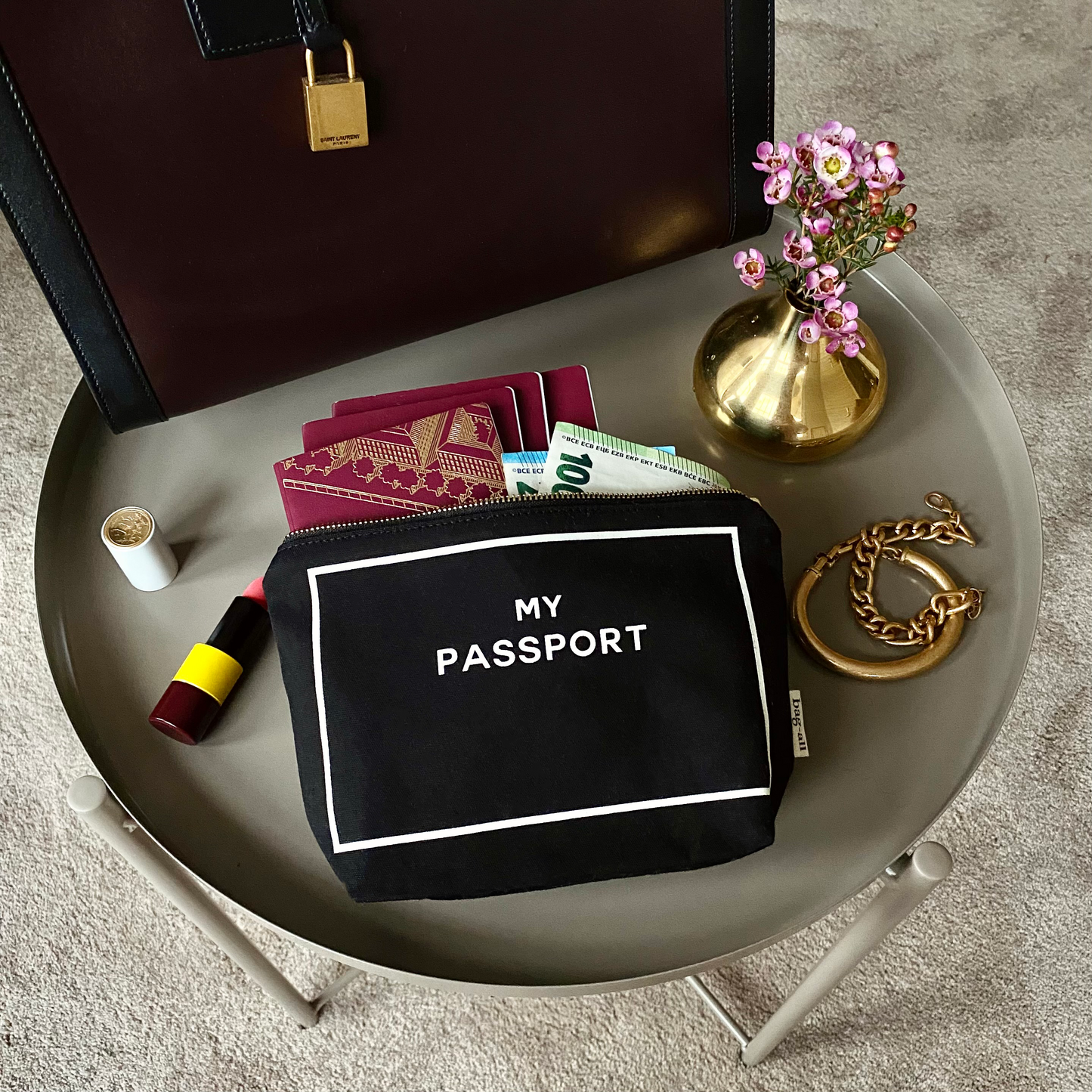 Bag-all Black Passport Pouch styled on gray tray with pink flowers, travel documents, and accessories. Sleek organizing solution for passports and cards.
