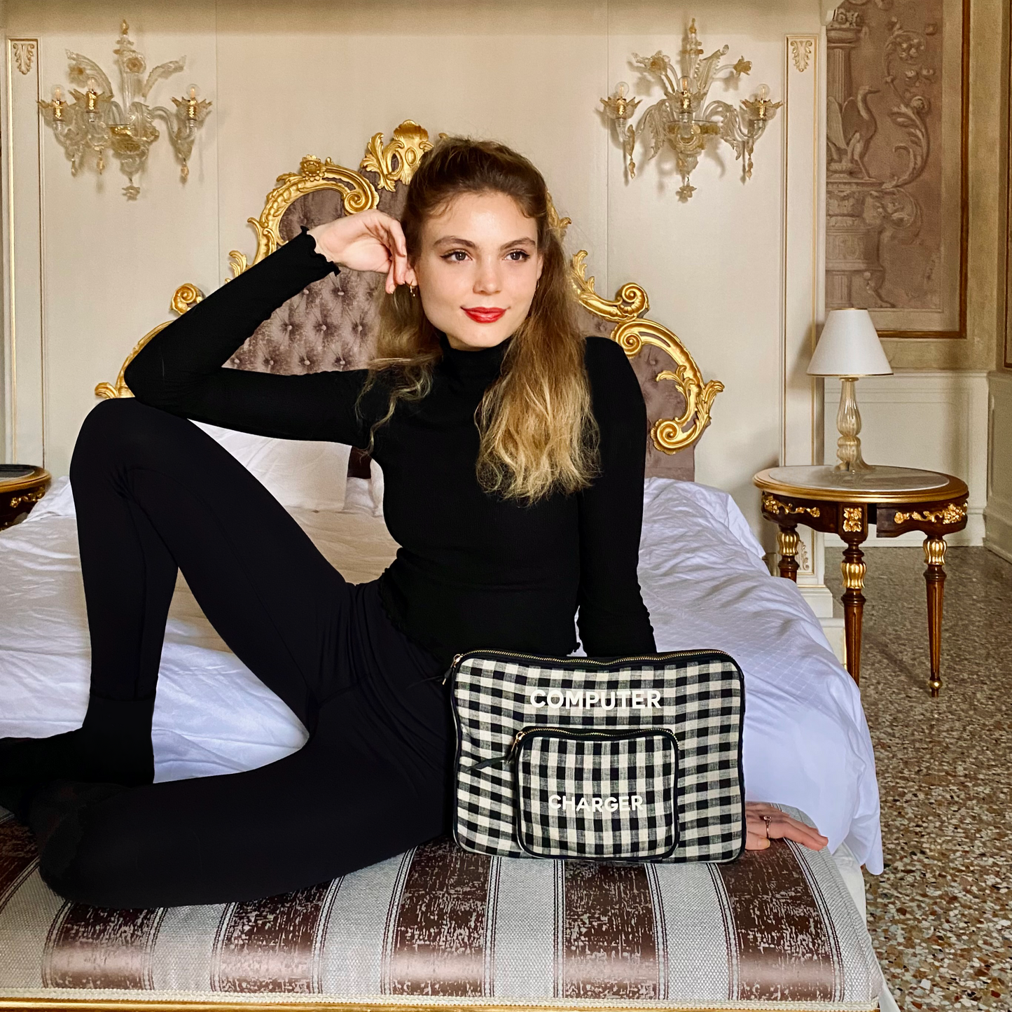 Bag-all black and white gingham laptop case with charger pocket shown on luxurious bed, featuring padded protection and gold-plated zipper for 13-inch devices