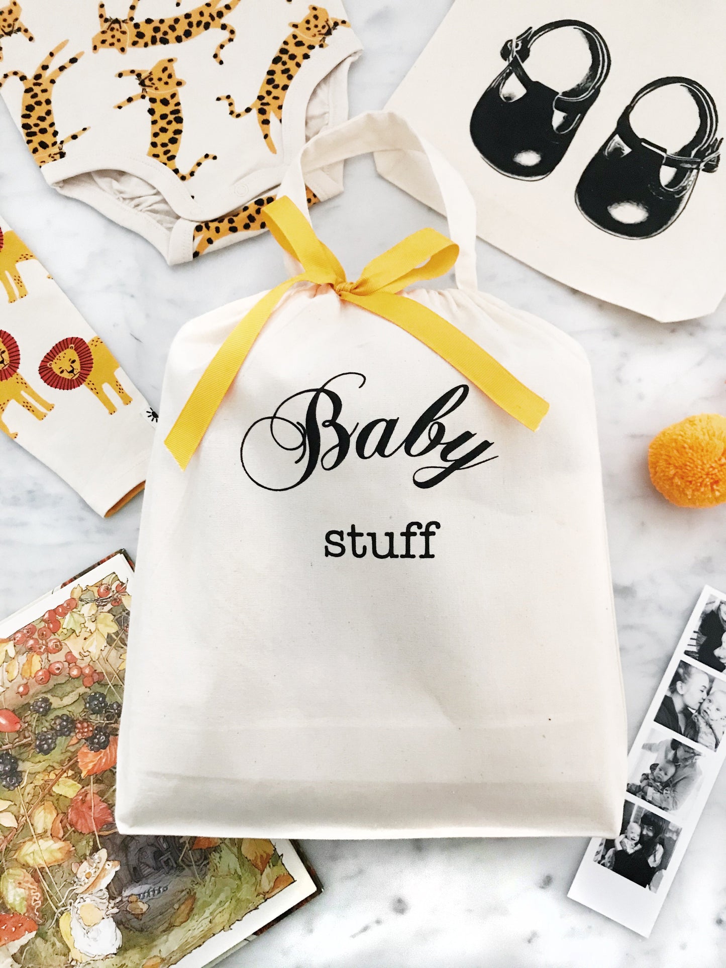 Bag-all Baby Stuff Organizing Bag with yellow ribbon, surrounded by animal print fabric, baby shoes, and vintage photos on marble surface