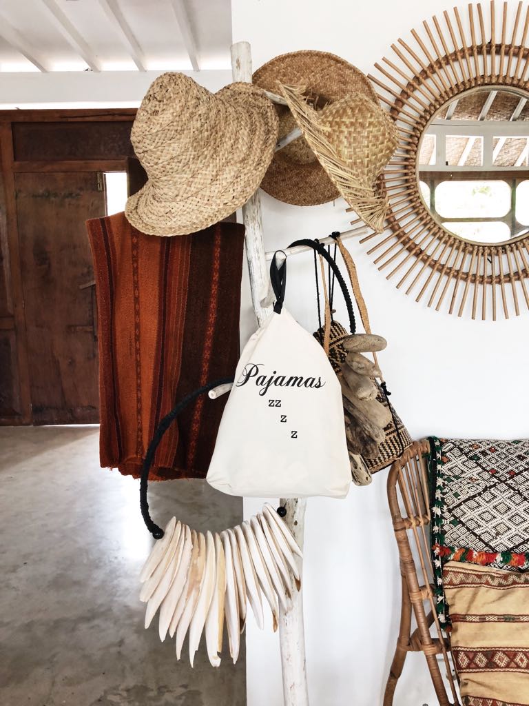 Bag-all Pajamas Zzzz Travel Bag in cream cotton with black text, hanging on wooden post alongside straw hats and rattan mirror, featuring drawstring closure and handle for convenient storage