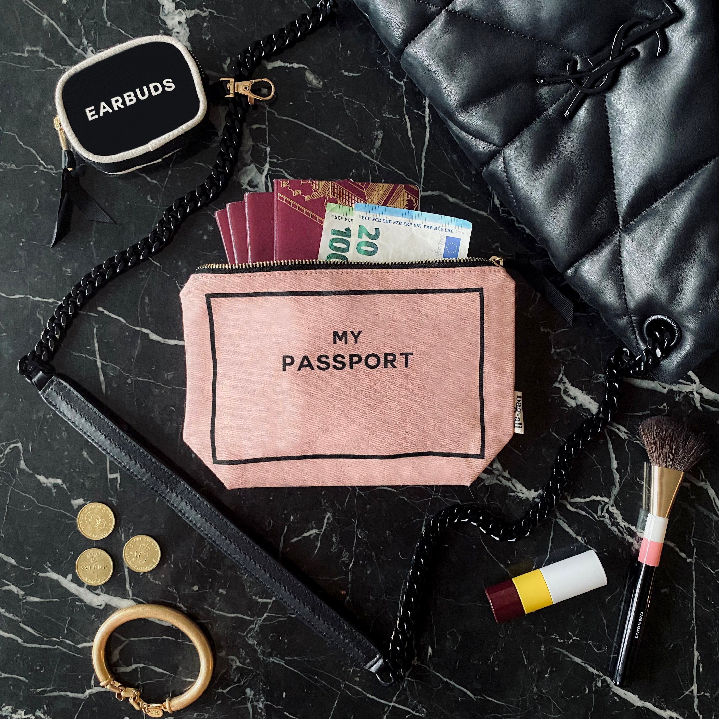 Bag-all Passport Pouch in Pink/Blush cotton with gold zipper and striped lining, styled on black marble surface with travel essentials including passports, currency, and accessories