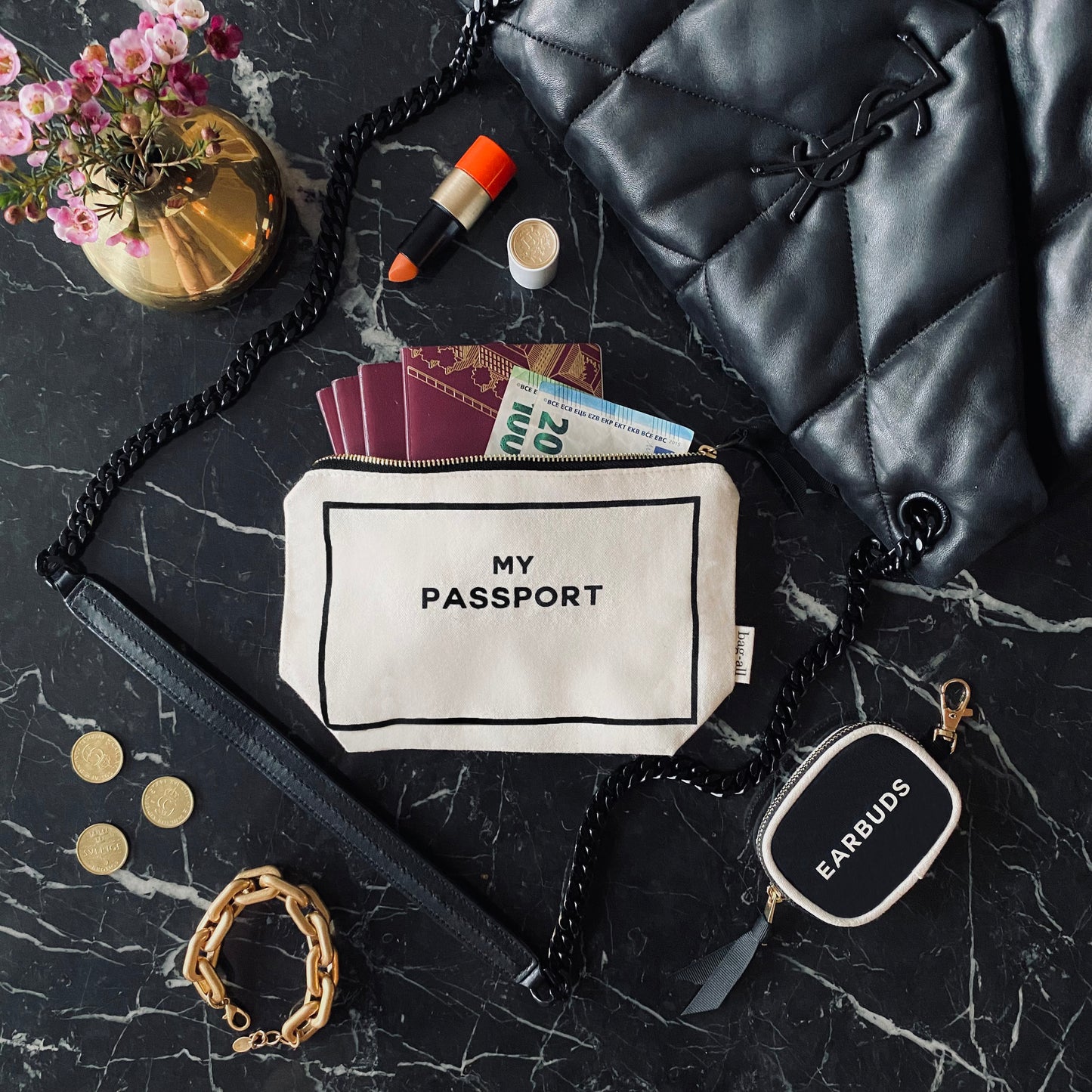 Bag-all Passport Pouch in cream cotton fabric with black text, shown open displaying passports and currency on marble surface, styled with black quilted bag and floral decor