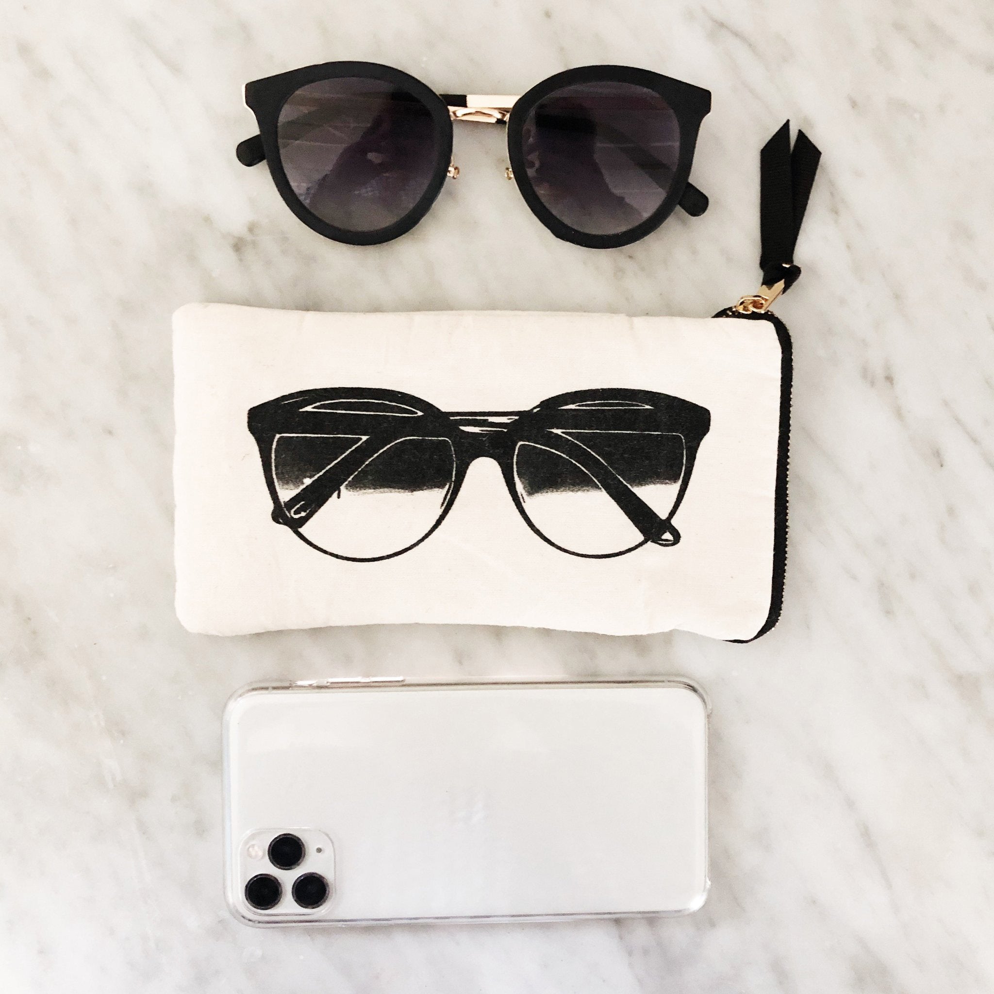 Bag-all Glasses Case with Outside Pocket in cream canvas featuring black sunglasses print, zippered compartment, and functional exterior pocket displayed with modern sunglasses and smartphone