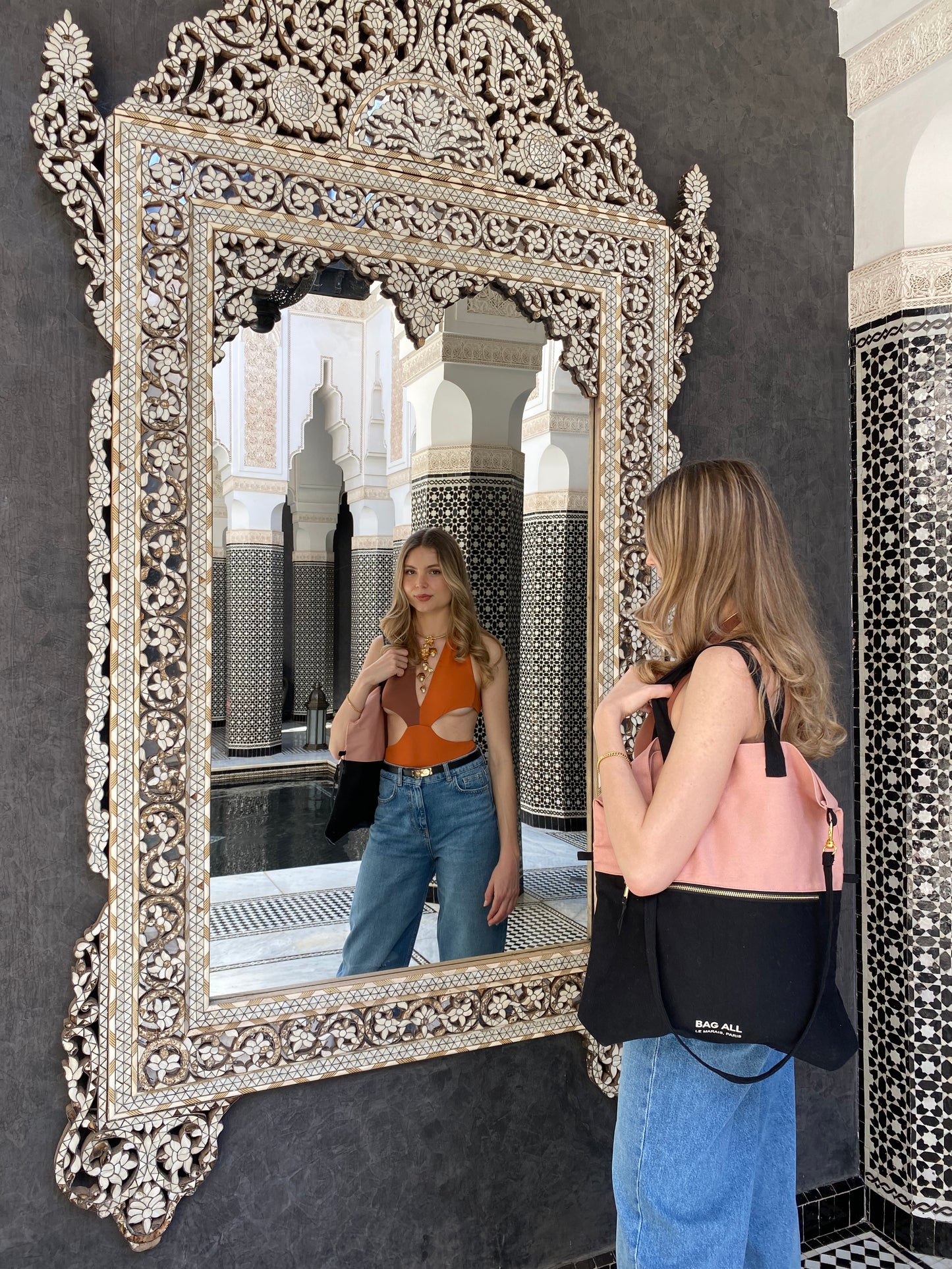 Bag-all stylish shopper wearing pink and black bag admiring ornate Moroccan mirror in traditional riad, showcasing intricate silver metalwork and Islamic architecture