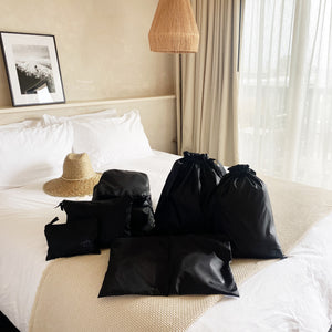 Bag-all Compression Cubes & Packing Bags Set in black displayed on white hotel bed with straw hat, featuring 8 recycled nylon travel organizers in various sizes for efficient packing