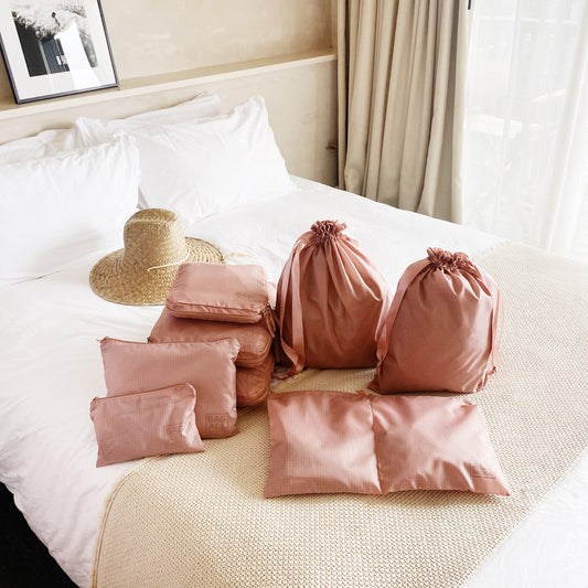 Bag-all Compression Cubes & Packing Bags Set in pink/blush, featuring 8 eco-friendly recycled nylon organizers displayed on white bedding with straw hat, perfect for sustainable travel