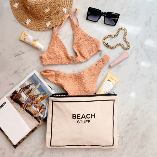 Bag-all Monogram Beach Pouch in cream canvas with black text, displayed with coral bikini, straw hat, sunglasses, and beach essentials on marble surface. Waterproof storage for swimwear and sunscreen.