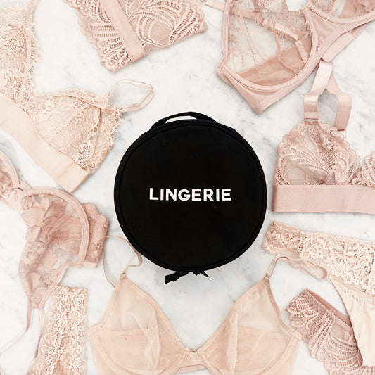 Bag-all Round Lingerie Case in black surrounded by blush pink lace bras and delicate undergarments on marble surface, featuring white LINGERIE text and ribbon handle