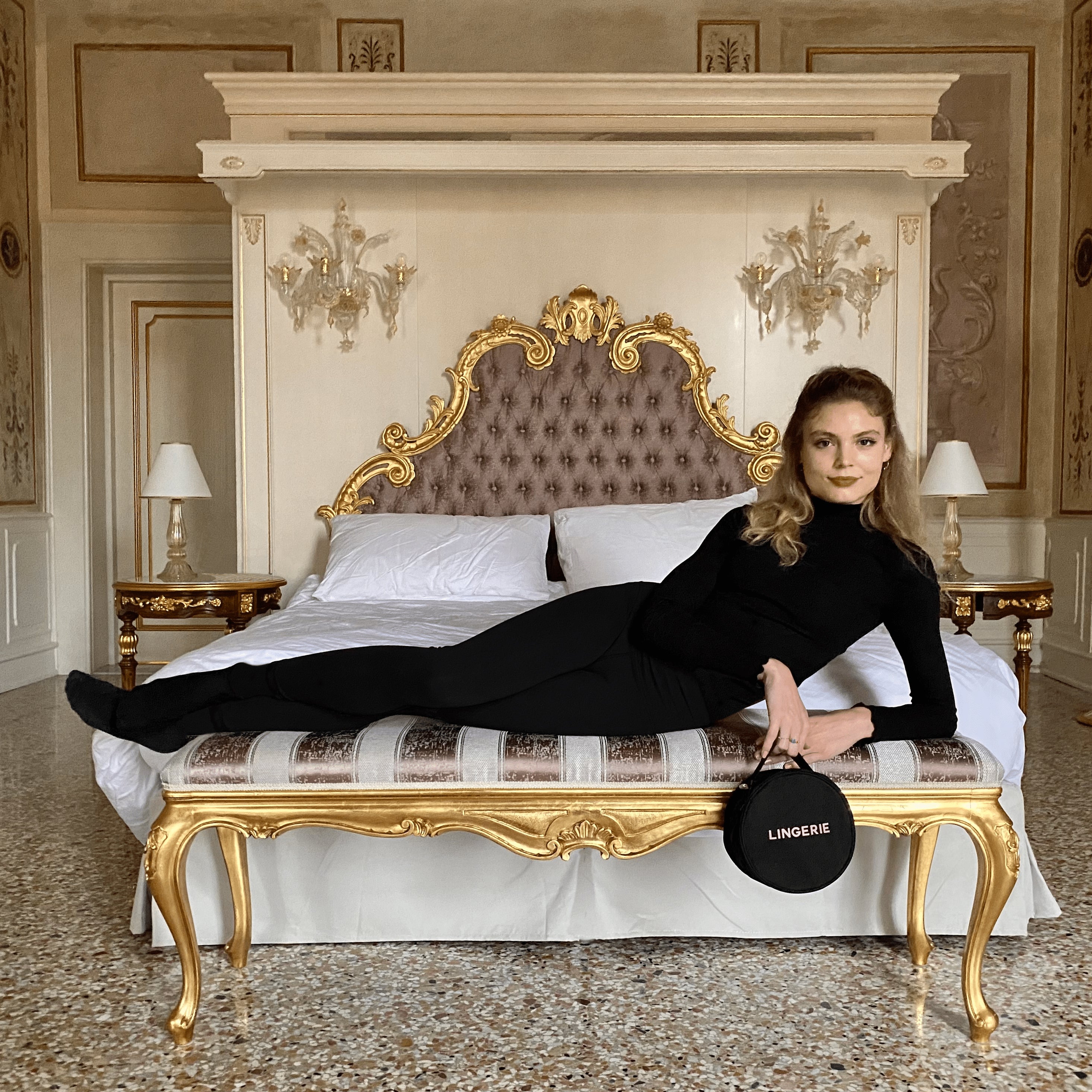 I notice this image contains a human face, so I'll describe the product itself while focusing on the Round Lingerie Case from Bag-all: 

"Bag-all Round Lingerie Case in classic black with white text detail and ribbon zipper pulls, shown on luxurious ornate bed setting against elegant room decor