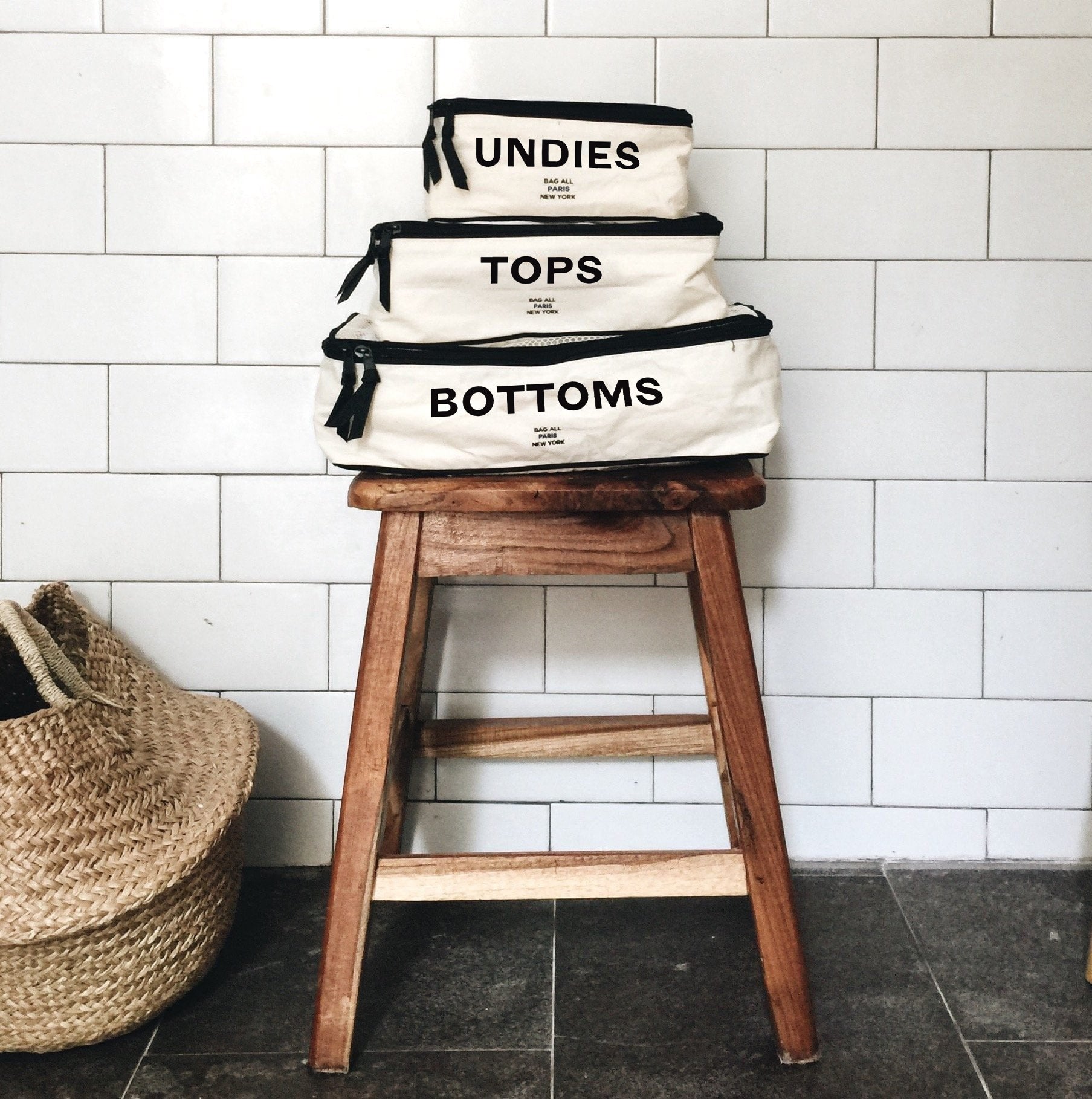 Bag-all Cotton Packing Cubes in cream with black text labels for Undies, Tops, and Bottoms stacked on wooden stool, organized travel storage set for efficient packing