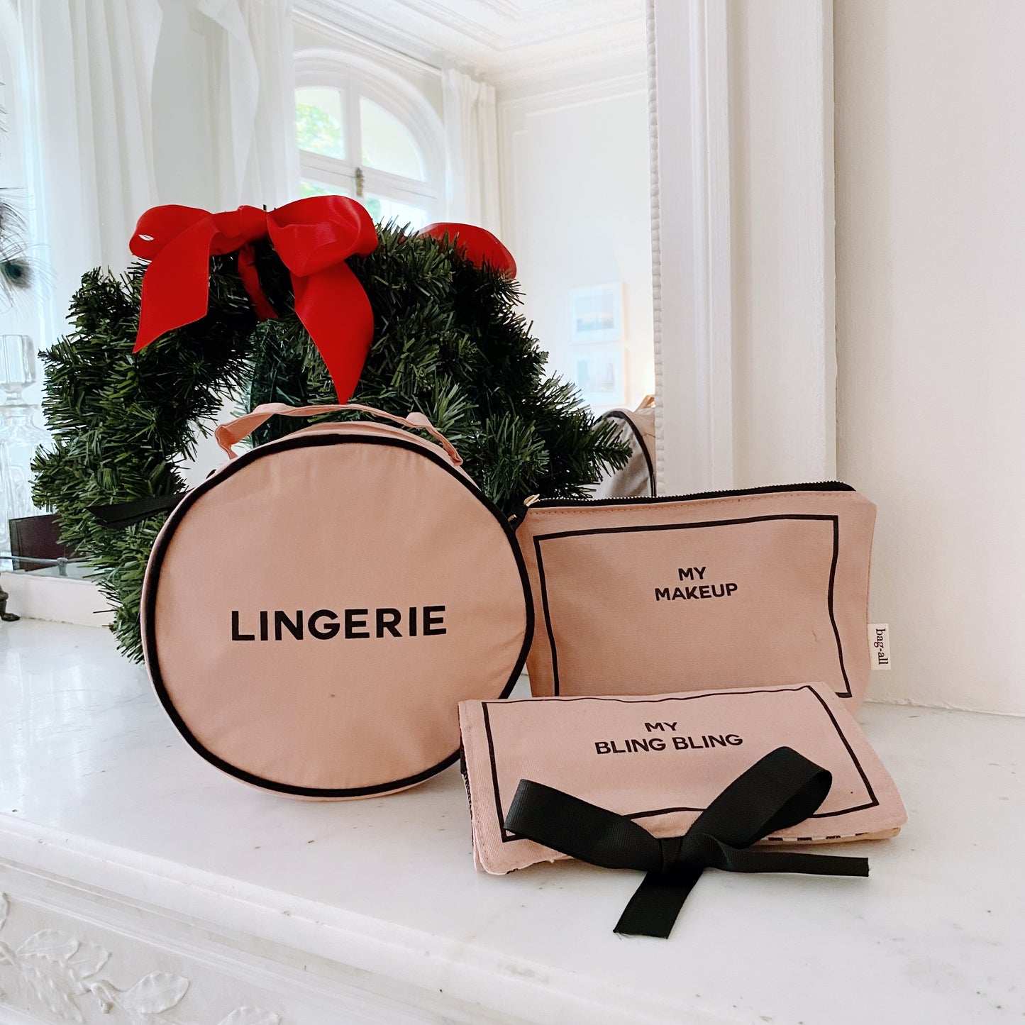 Pink and blush Bag-all 3-pack set with round lingerie case, makeup case, and jewelry pouch with black bow near Christmas wreath