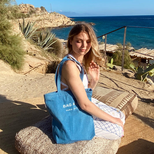 Bag-all Paris Le Marais denim tote with zipper showcased at scenic coastal location, featuring blue fabric, white text print, and practical inside pocket for everyday use.