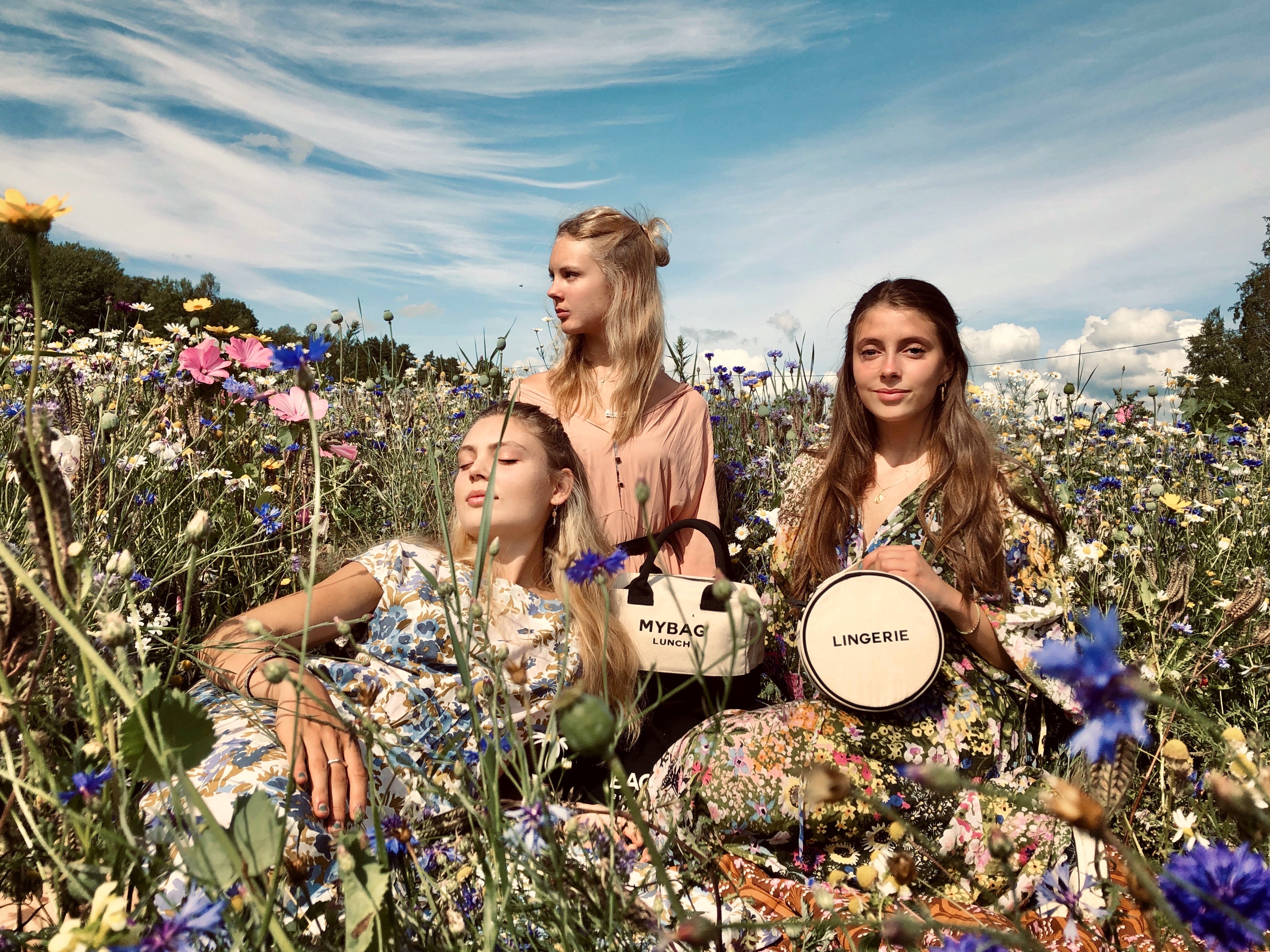 Three women in floral dresses sitting in a wildflower field holding personalized cotton travel bags and a round lingerie case by Bag-all.