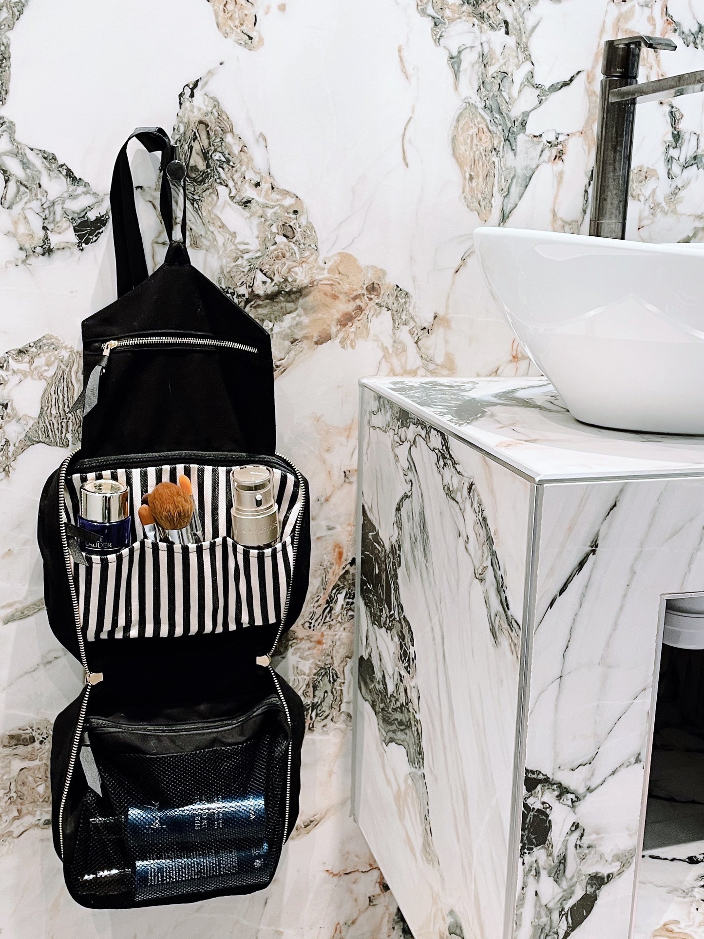 Bag-all black hanging toiletry organizer open with grooming products inside on marble bathroom wall background