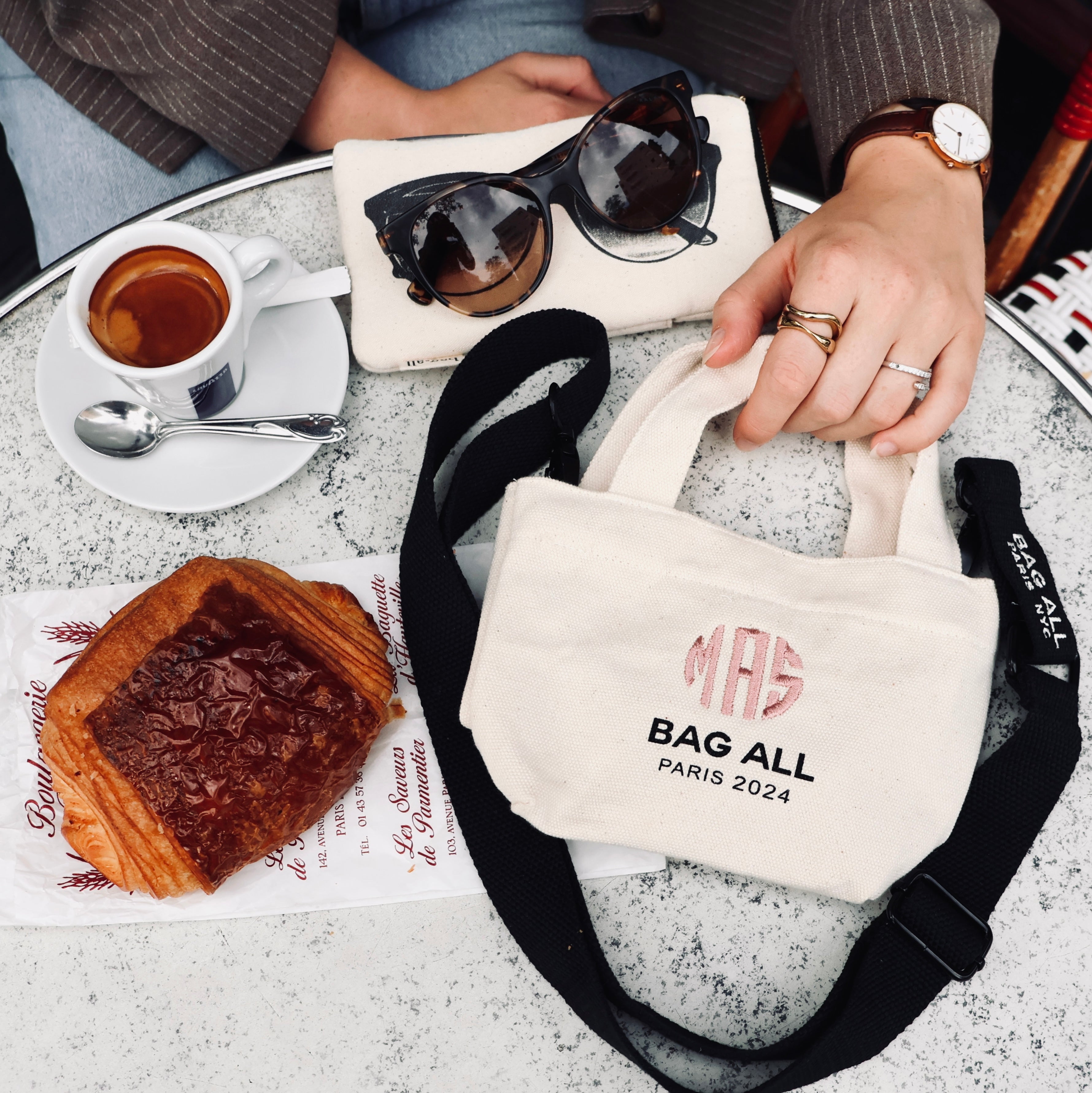 Bag-all Mini Tote Bag with monogram shown at cafe table with croissant and espresso, featuring cream canvas material, black strap, and Paris 2024 branding