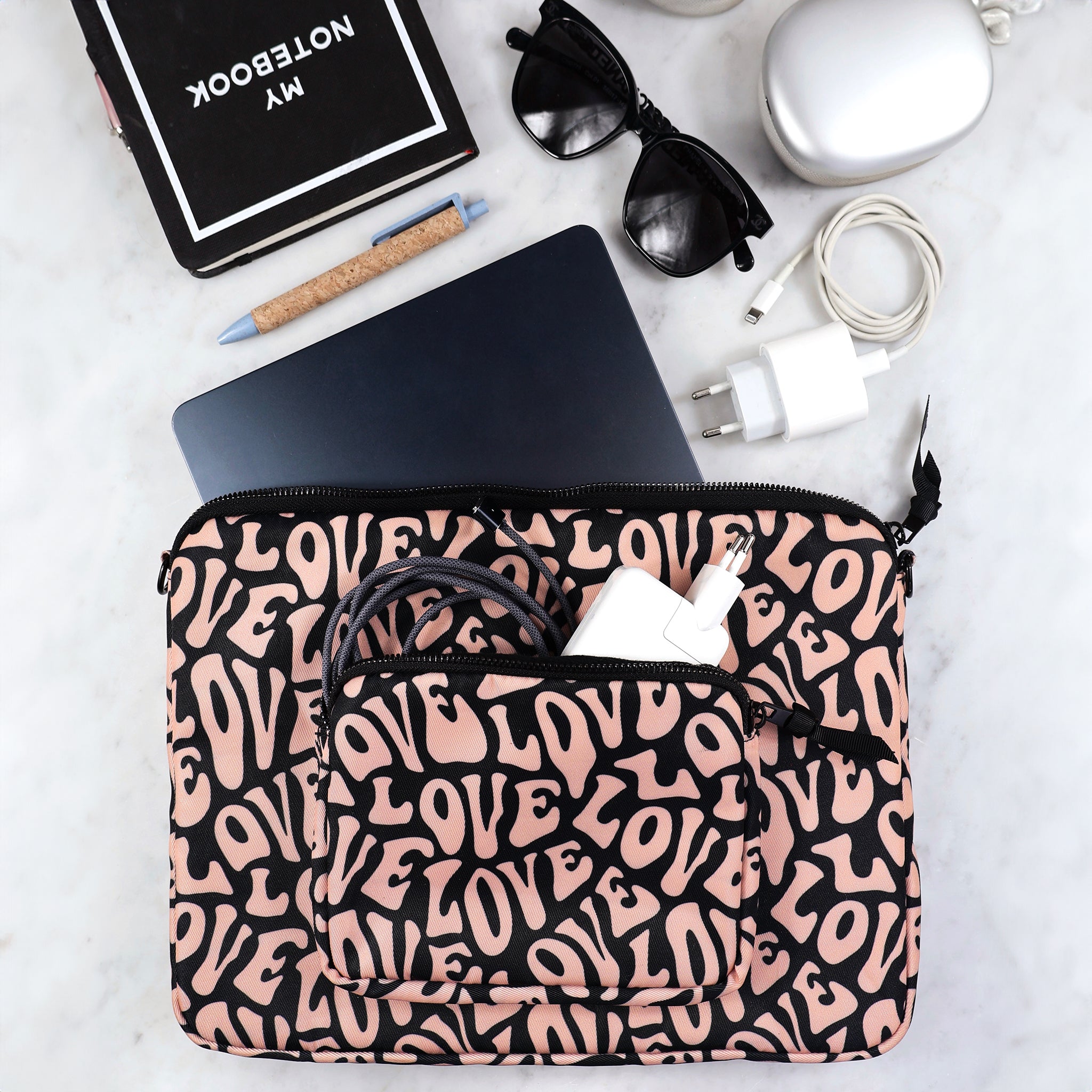 Bag-all padded laptop case with pink love pattern and charger pocket, featuring black zipper and eco-friendly design, styled with sunglasses and accessories on marble surface
