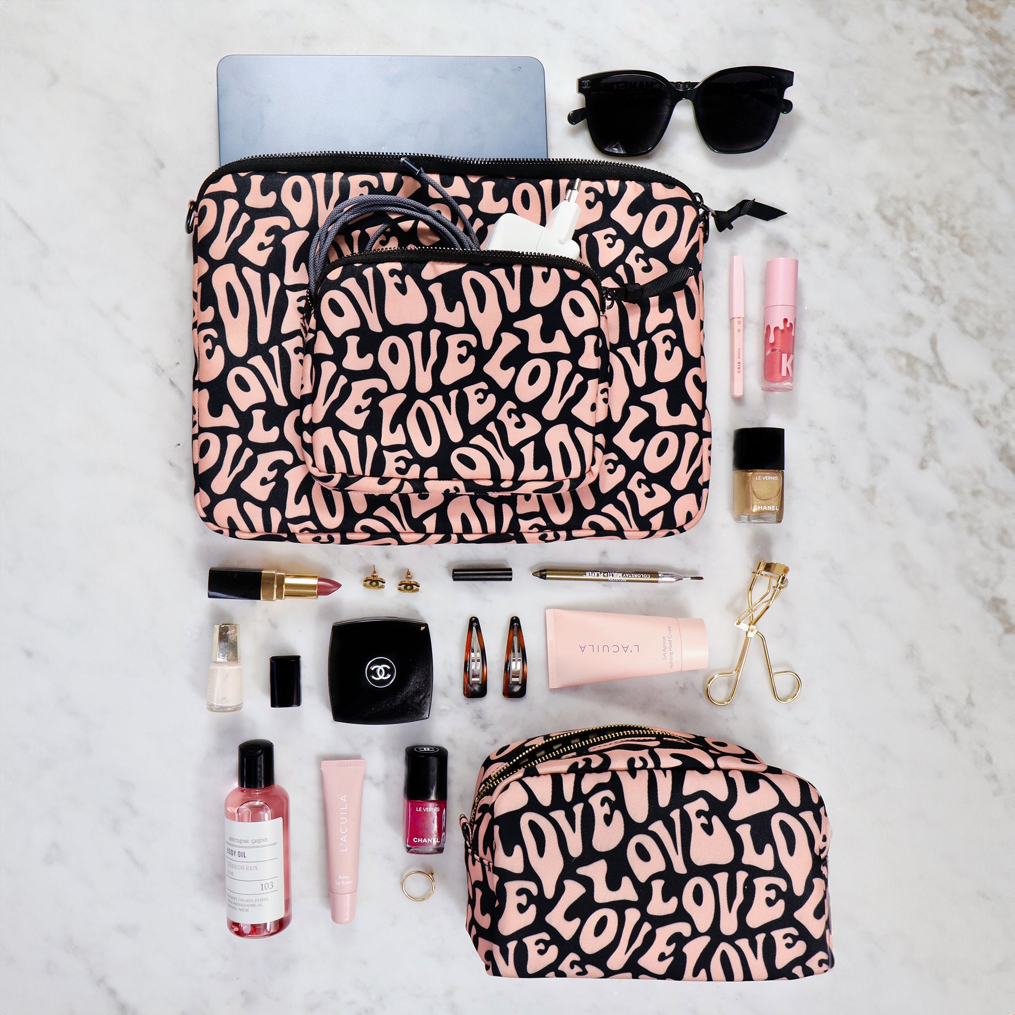 Bag-all Toiletry Pouch with pink and black love pattern displayed with beauty essentials, sunglasses, and cosmetics on marble surface, featuring eco-friendly recycled material construction and water-resistant interior
