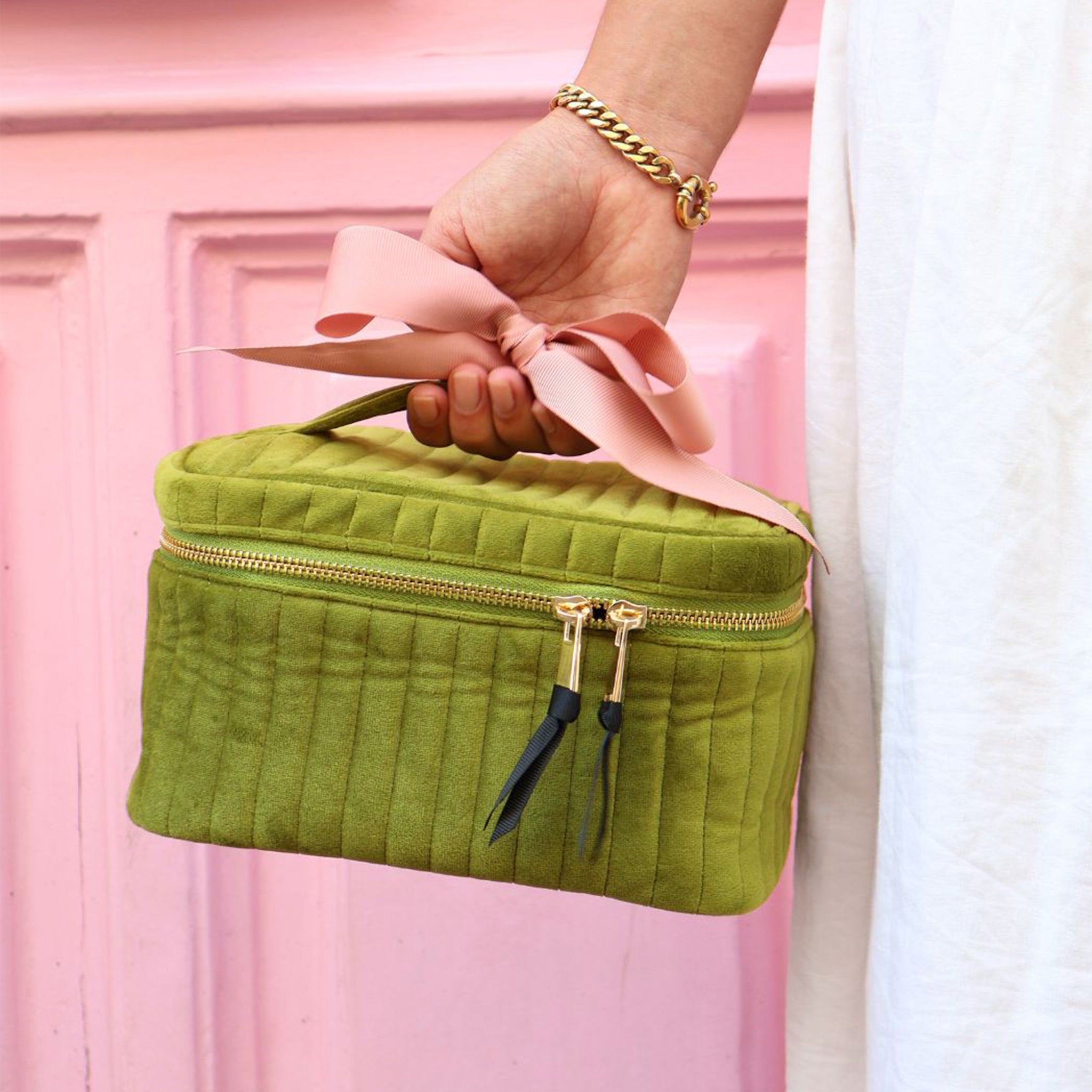 Green quilted eco velvet makeup case with gold zipper and pink ribbon handle by Bag-all held against pink background
