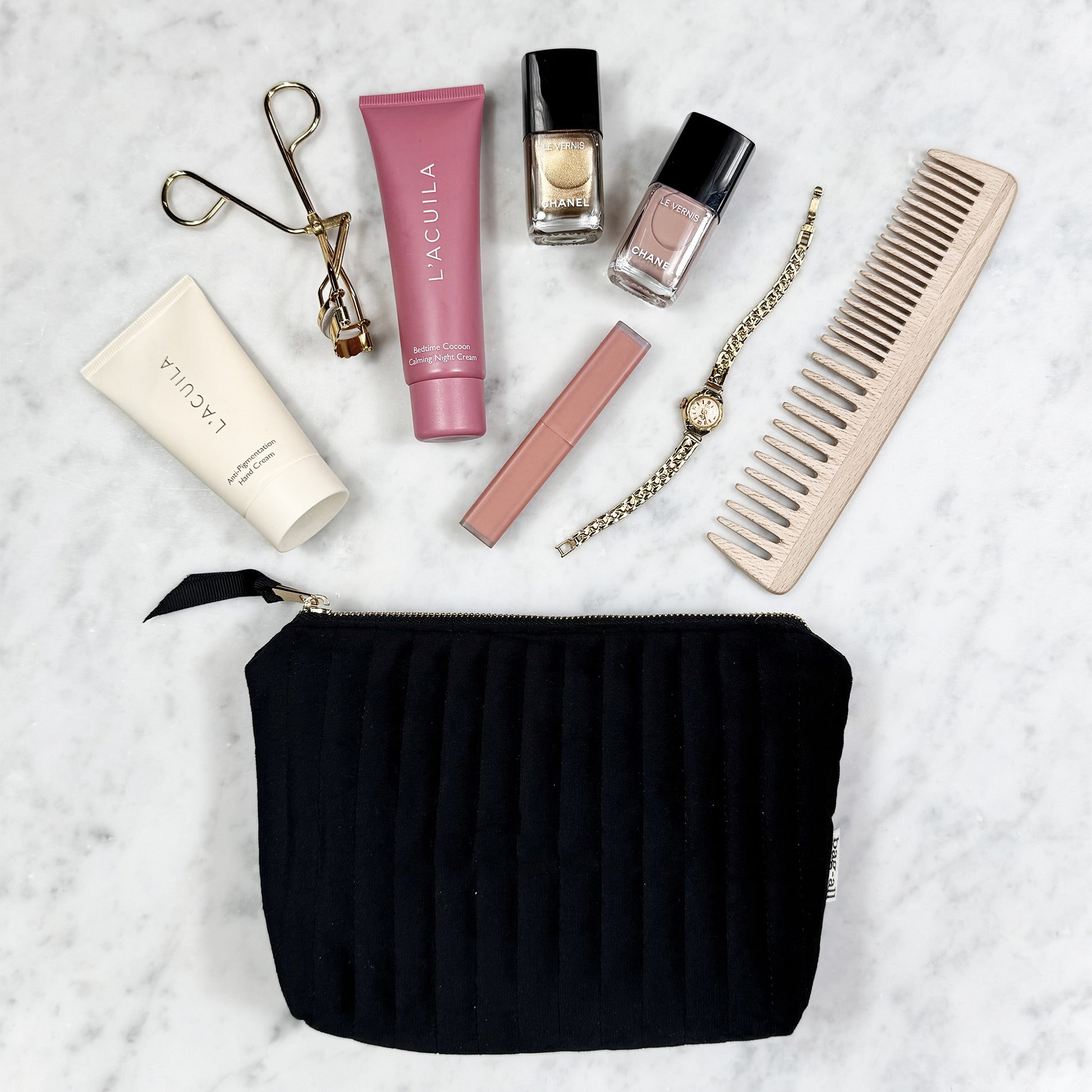 Black eco-velvet Bag-all pouch with makeup, nail polish, gold watch, comb, and eyelash curler on marble background