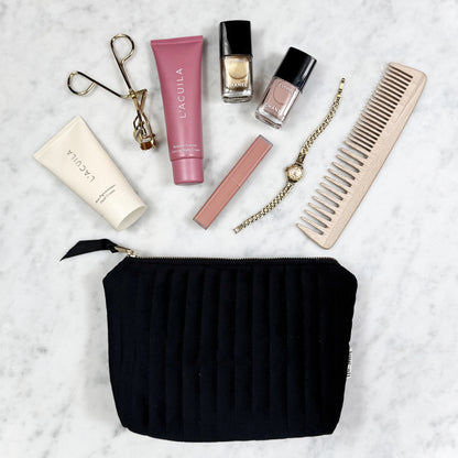 Black eco-velvet Bag-all pouch with makeup, nail polish, gold watch, comb, and eyelash curler on marble background