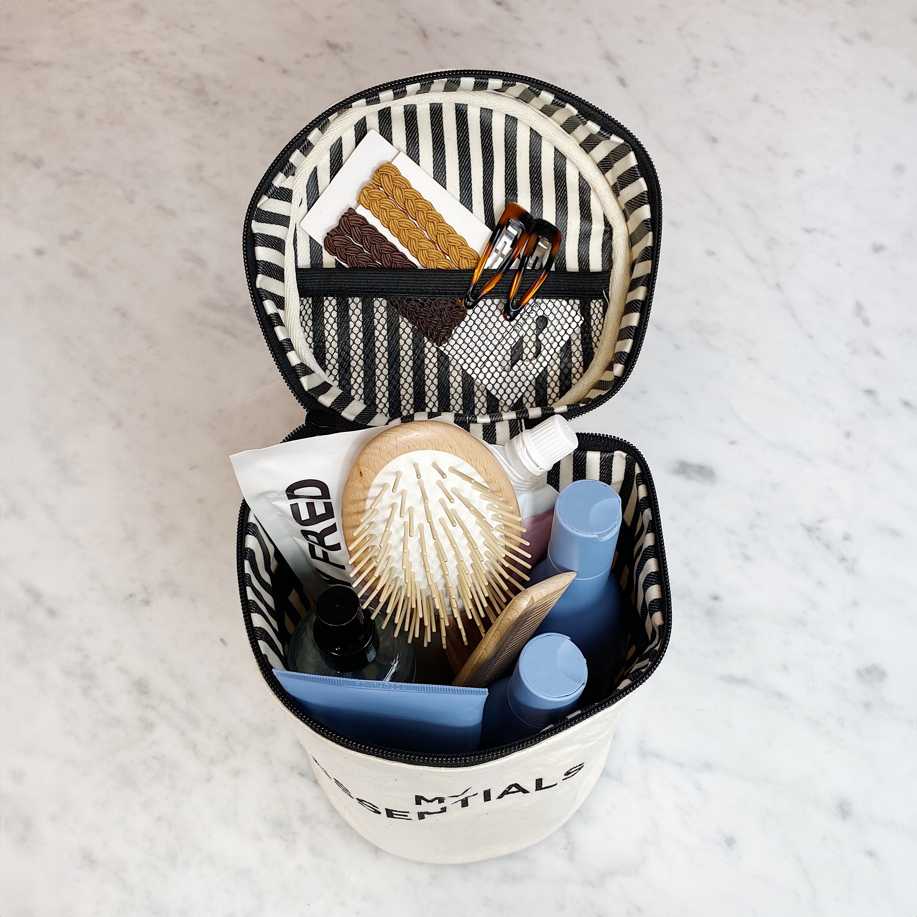 Bag-all Roomy Travel Essentials Case in cream with striped interior, shown open displaying hairbrush, cosmetics and toiletries organized in compartments with waterproof lining