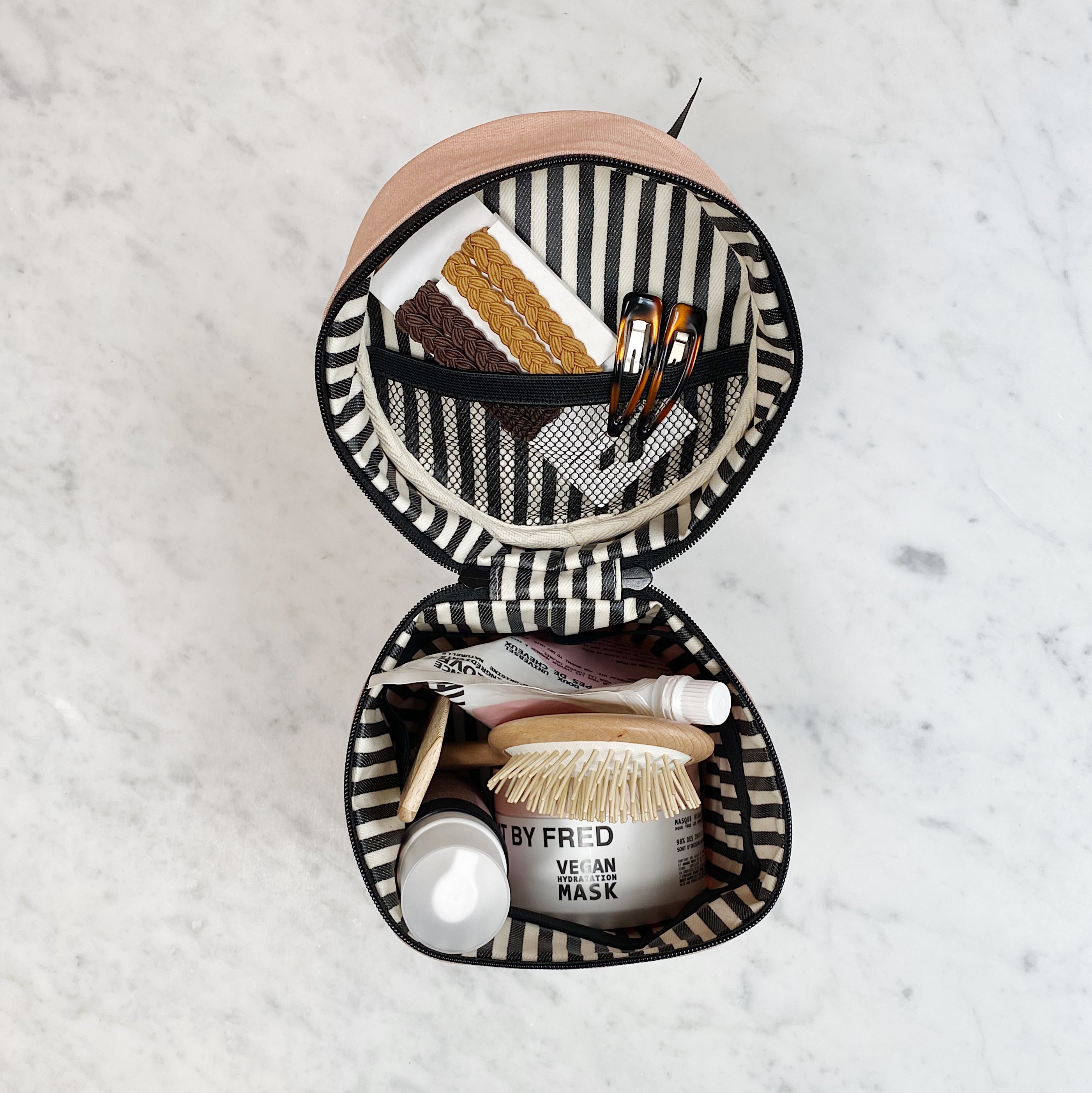 Bag-all Round Travel Essentials Case in Pink/Blush featuring striped interior, waterproof lining, elastic holders, and mesh pocket, displayed open with beauty products and brushes organized inside on marble surface