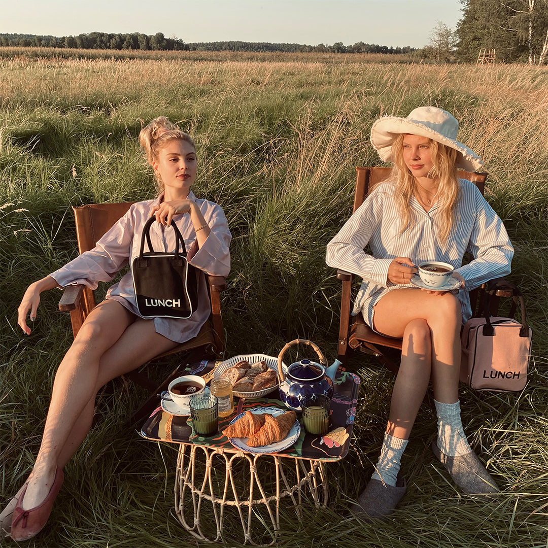 Two women sitting outdoors in grassy field enjoying tea and pastries with Bag-all personalized cotton lunch bags for stylish travel organization