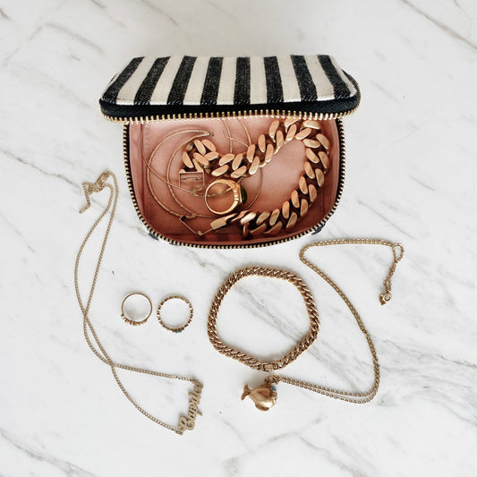 Bag-all Jewelry/Trinket Box in black and white stripes, shown open displaying gold jewelry pieces including chains, rings, and bracelets on marble surface. Compact, travel-friendly design with gold zipper.