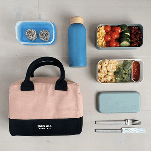 Bag-all Stylish Monogram Lunch Box in pink/blush with black trim, shown with healthy meal prep containers, blue water bottle and utensils in a minimalist flatlay arrangement