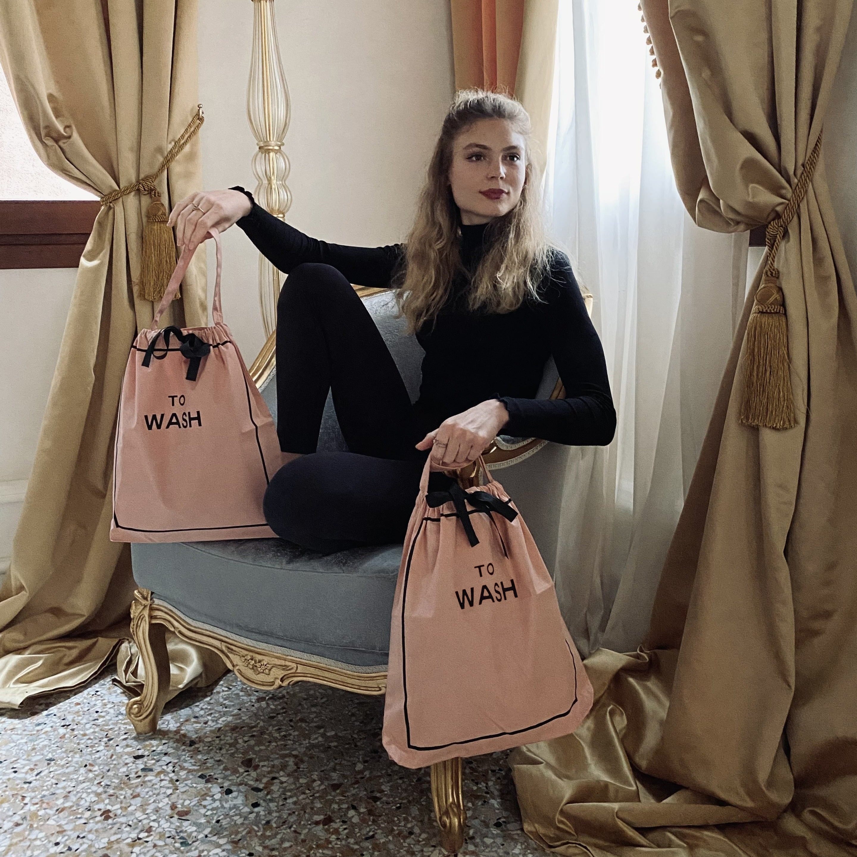 Woman holding two pink Bag-all laundry organizers sitting on vintage chair in elegant room with curtains