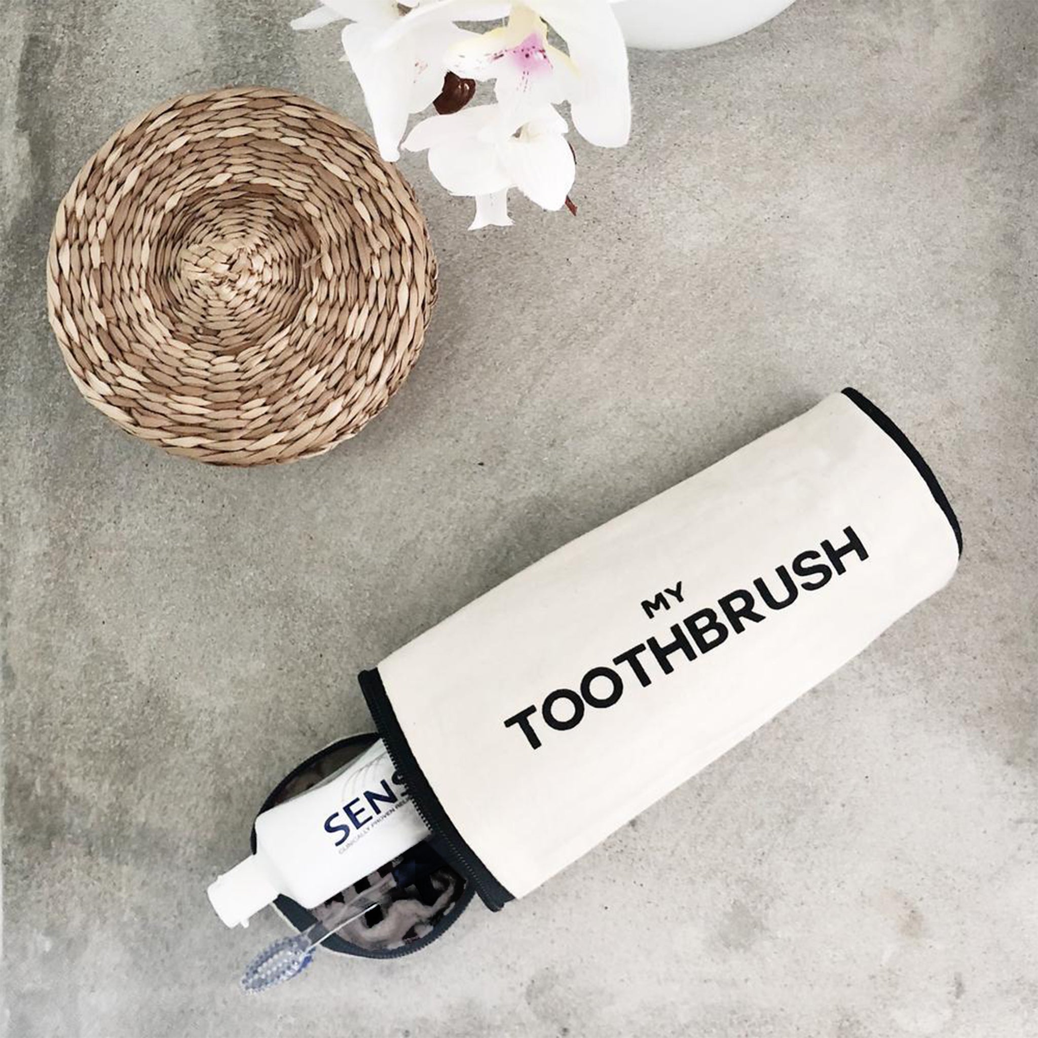 Bag-all My Toothbrush travel case in cream cotton with black text, displayed on grey surface near woven coaster and white orchid, part of Toiletry Travel Gift Set Deal 4-Pack