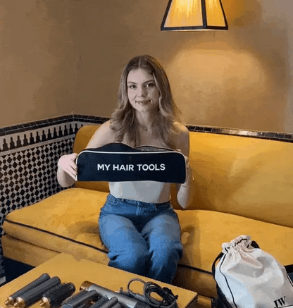 Woman sitting on yellow couch holding black Bag-all hair tools organizer pouch in cozy room with tiled wall background
